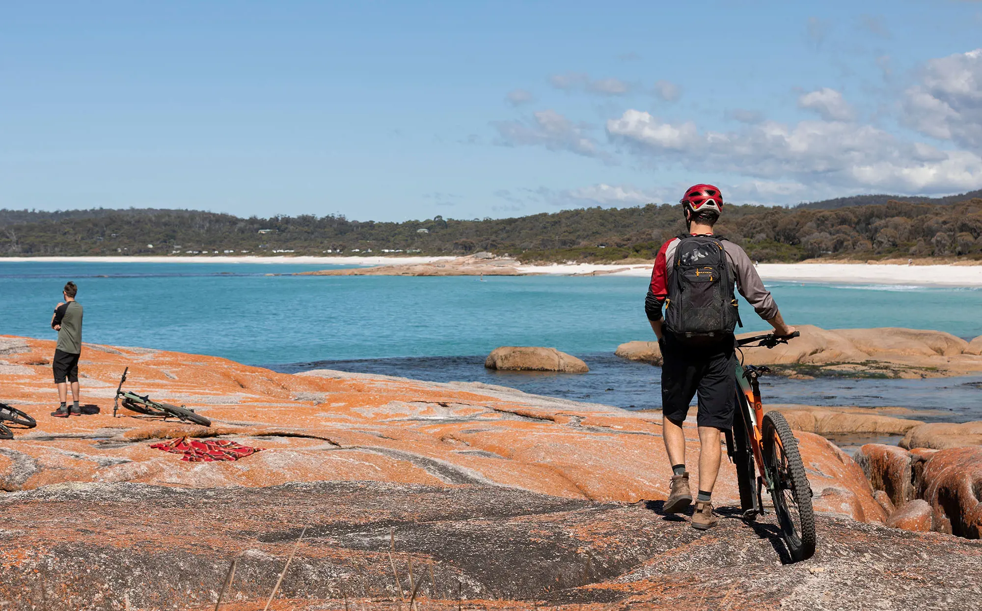 A mountain biker walks his bike across wide, flat rocks next to the clear blue waters of a beach.