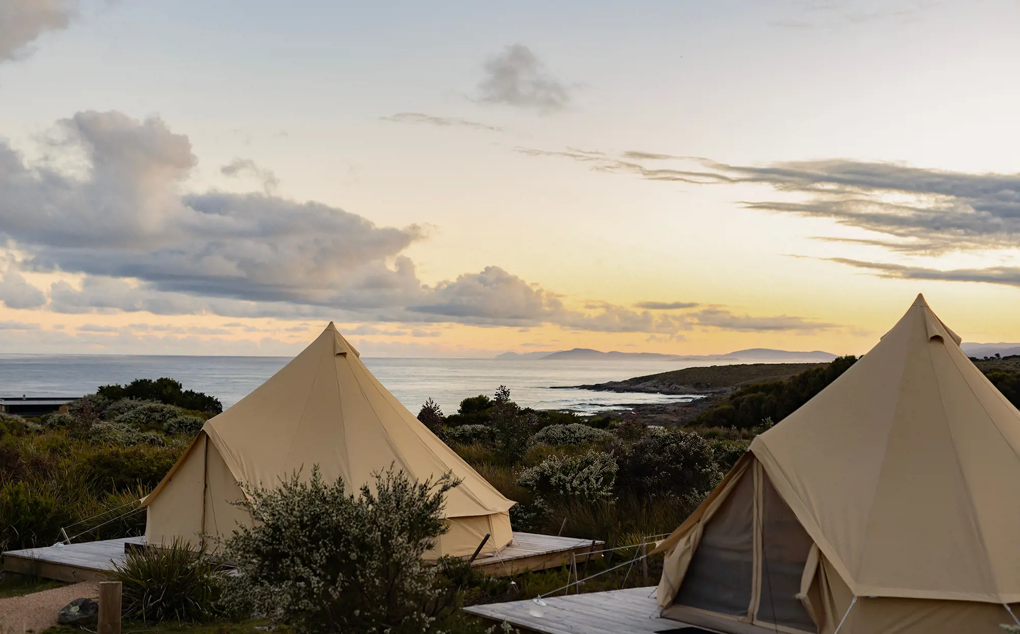 Two beige canvas tents are set up on wooden platforms in a coastal landscape at sunset. The ocean and distant hills are visible under a partly cloudy sky.