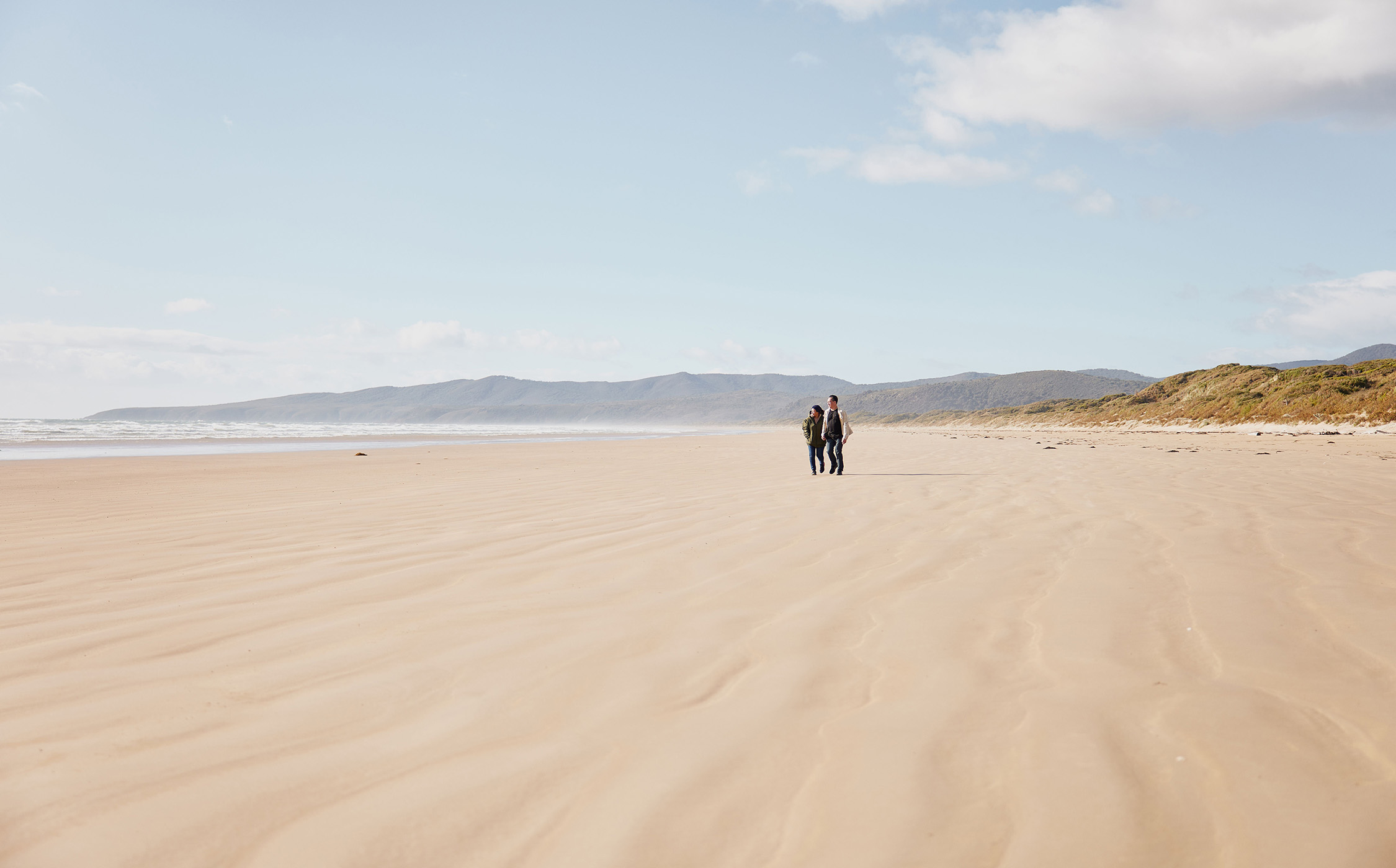 Two people walk in the distance on the wide expanse of a sandy beach. Mountains rise in the far distance.