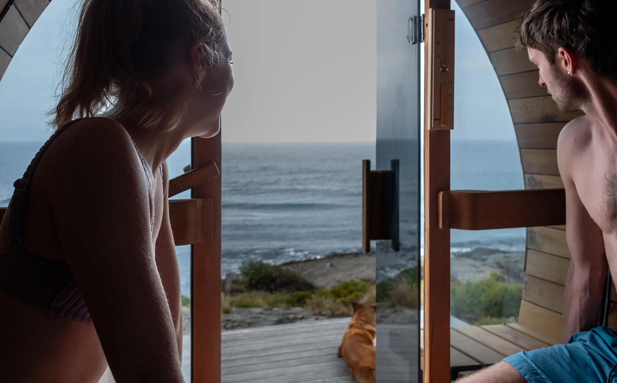 Two people in swimsuits sit inside of a sauna with glass doors and windows, on a wooden deck. The door is open and they are looking out to the ocean beyond.