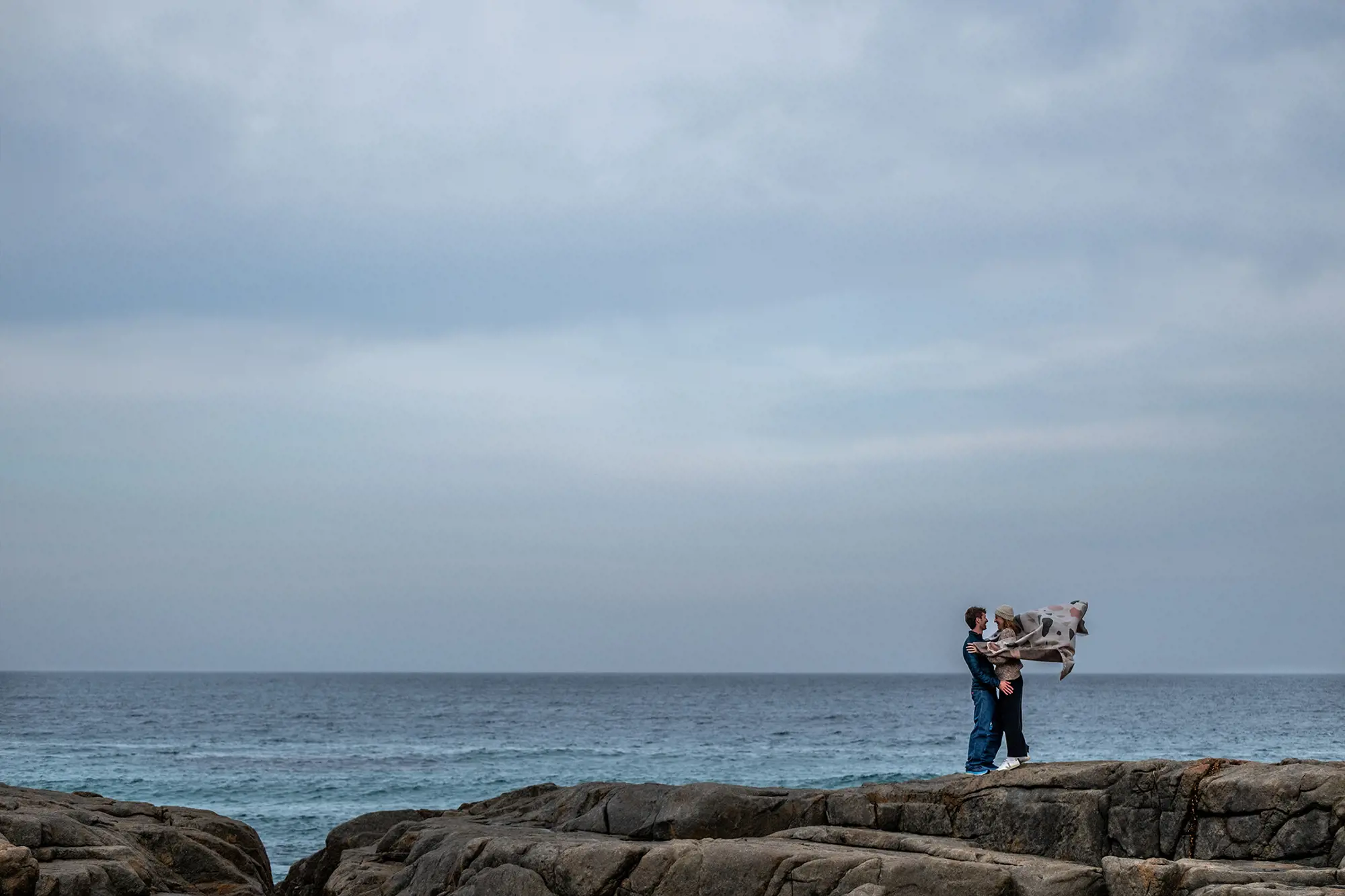Two people embrace, standing on rocks above an ocean that stretches out to the horizon. Her cardigan is billowing out behind her from the wind.