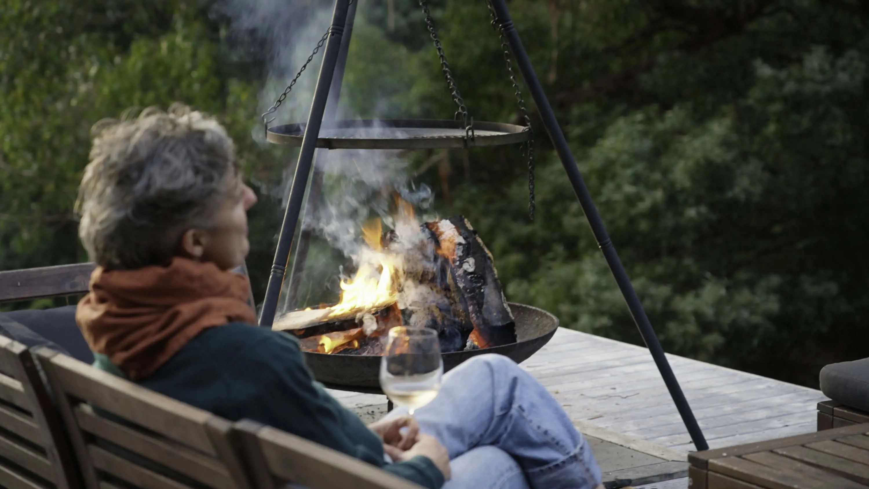 A person holding a glass of white wine sits on a bench beside a small hanging firepit, where logs are blazing.