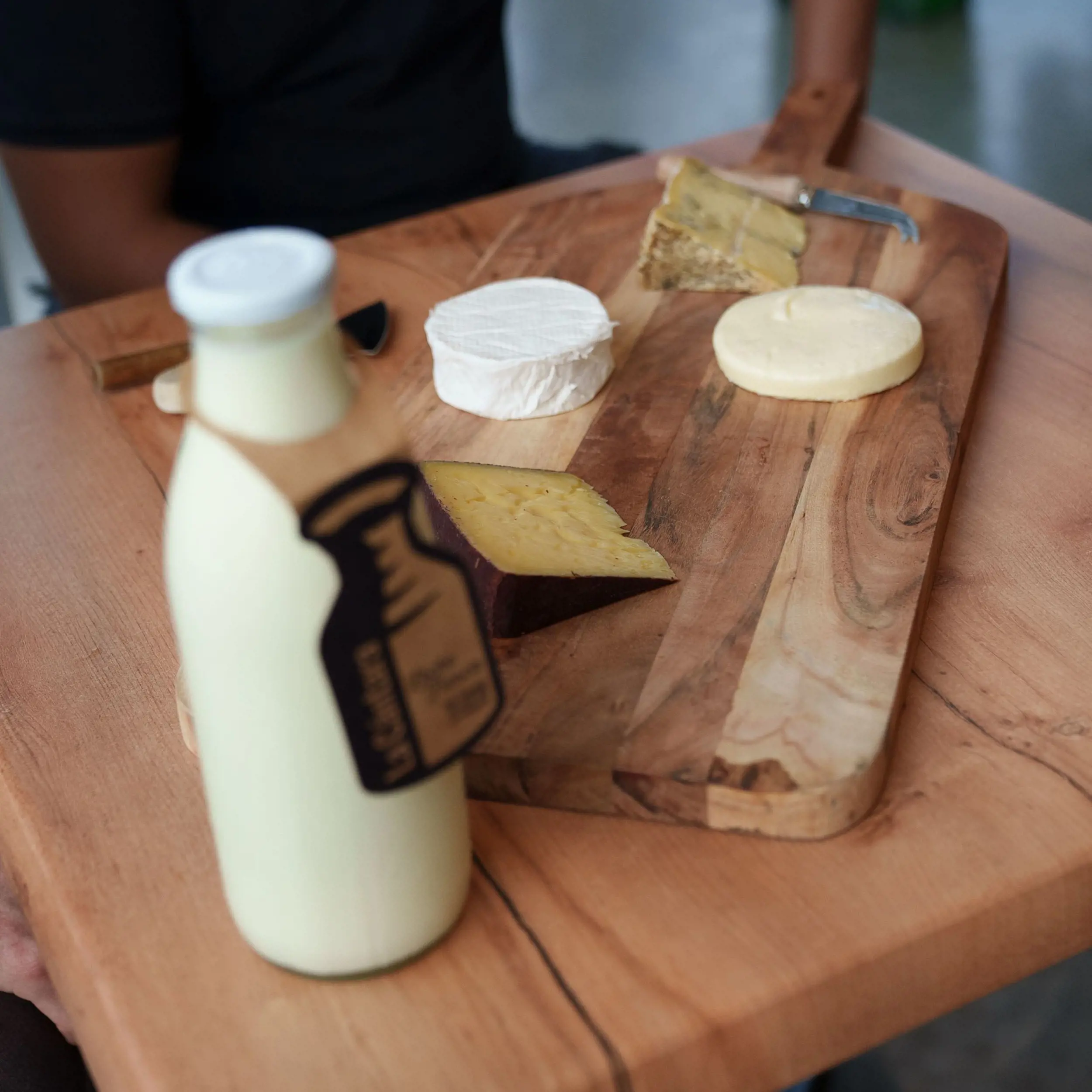 Four different pieces of cheese, arranged on a wooden board with several cheese knives. Next to the board is a 1 litre glass bottle full of milk.