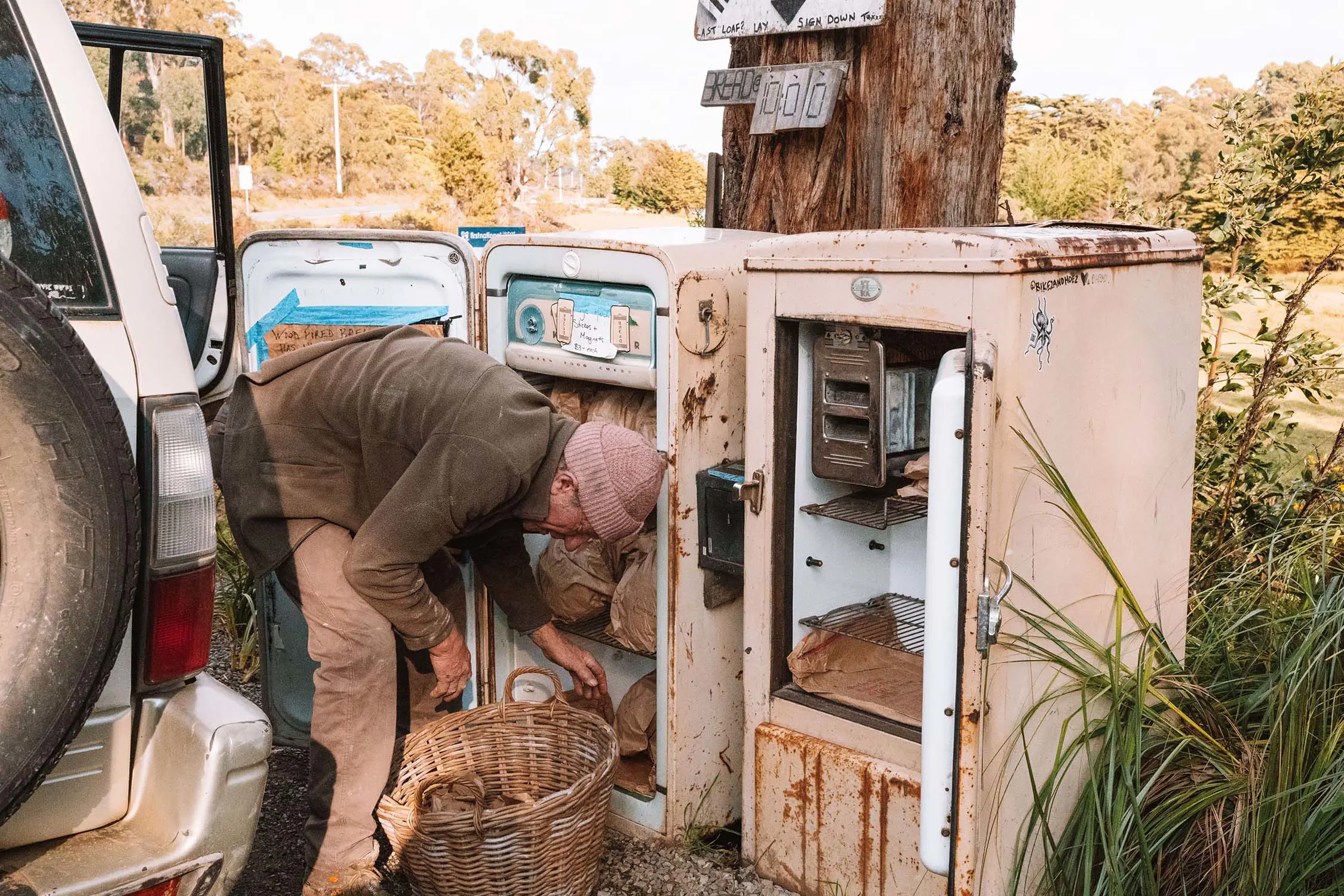 Next to a row of old, weathered fridges standing on the side of a road under a tree, a man is bending over a basket, filling the shelves with freshly baked loaves of bread.