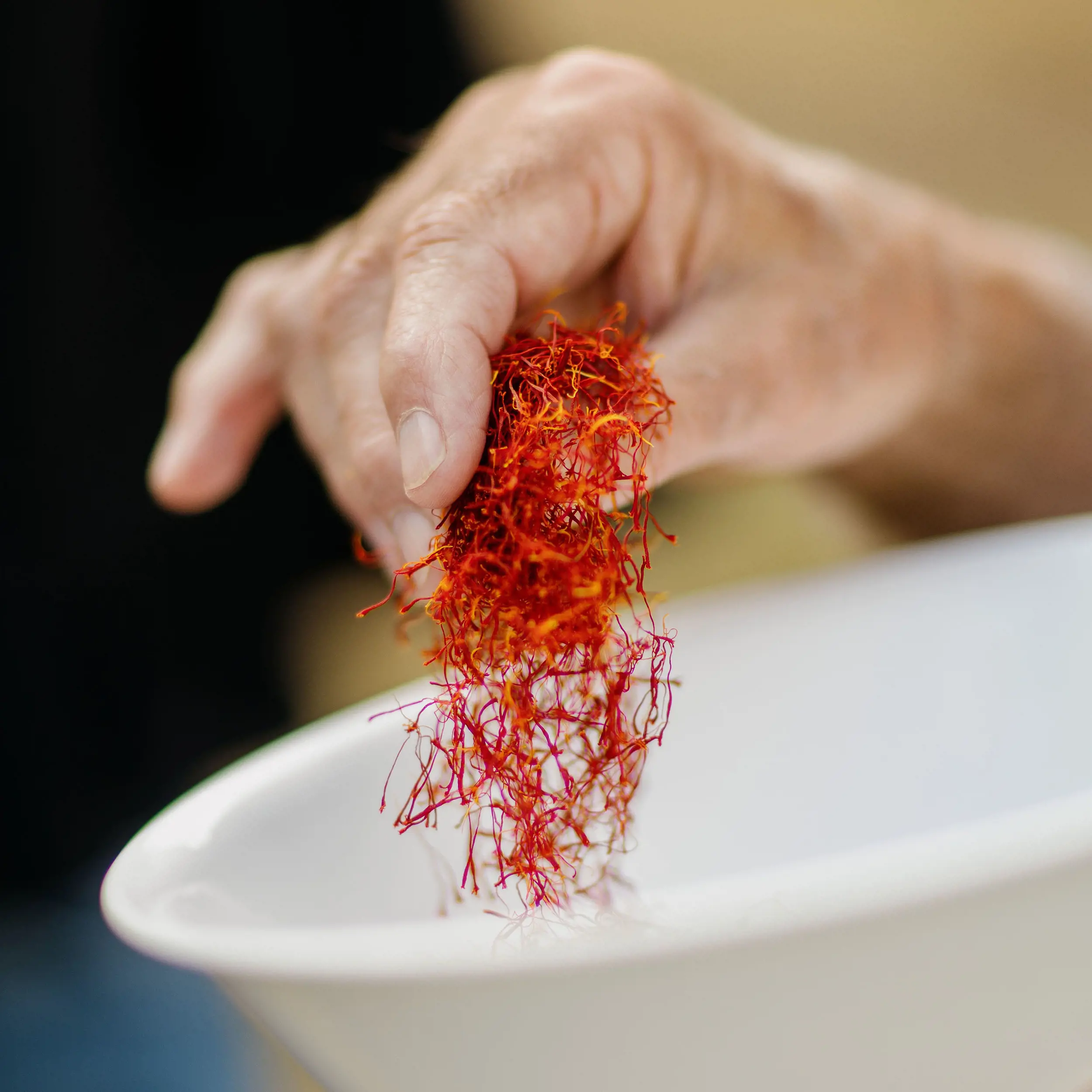 A close-up of a person's hand picking up a large pinch of bright red saffron from a white bowl.