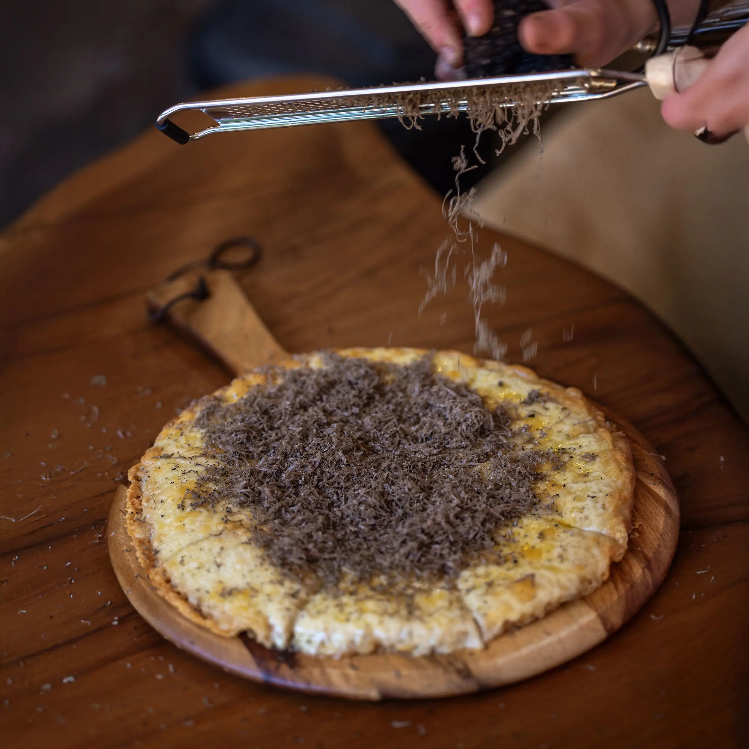 A fresh, cheesy woodfired pizza on a wooden paddle is being loaded with truffle shavings, being grated onto the pizza.