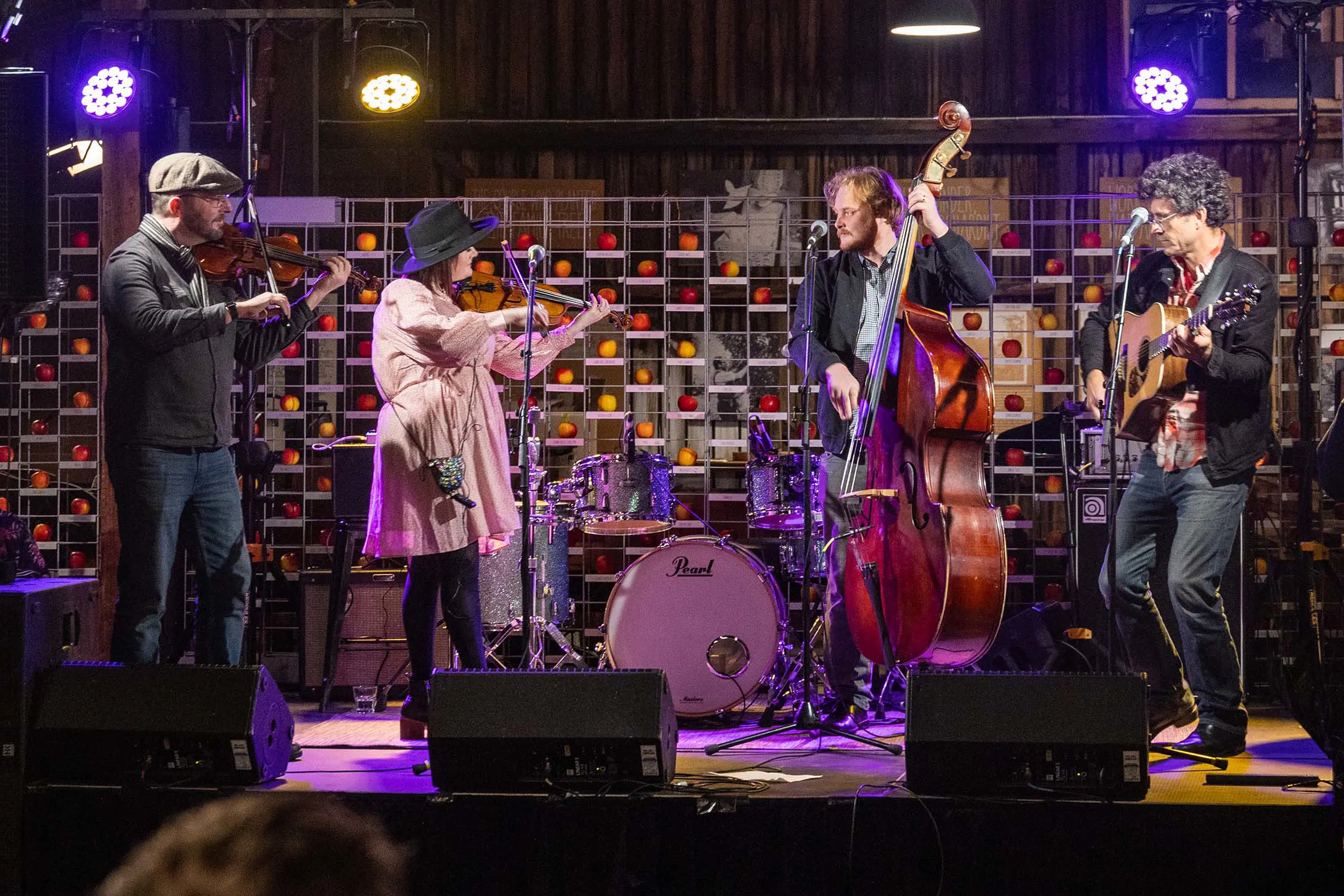 A quartet of folk musicians play on a small stage, in front of a wall decorated with a huge variety of kinds of apples.