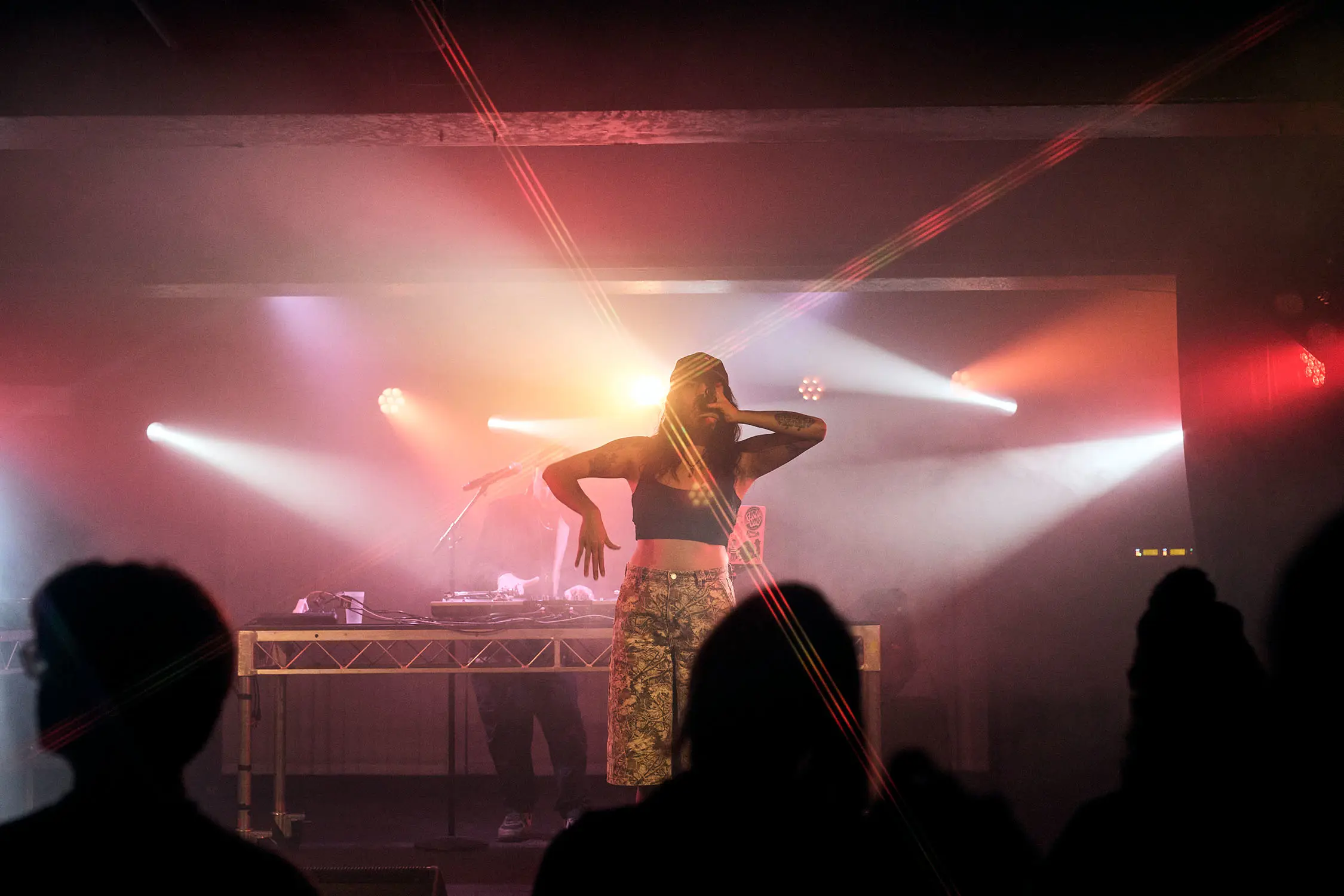 A woman performs on a hazy stage in a small underground bar, while a DJ spins behind her. The silhouettes of the audience are visible, and the lights reflect to create beams across the image.