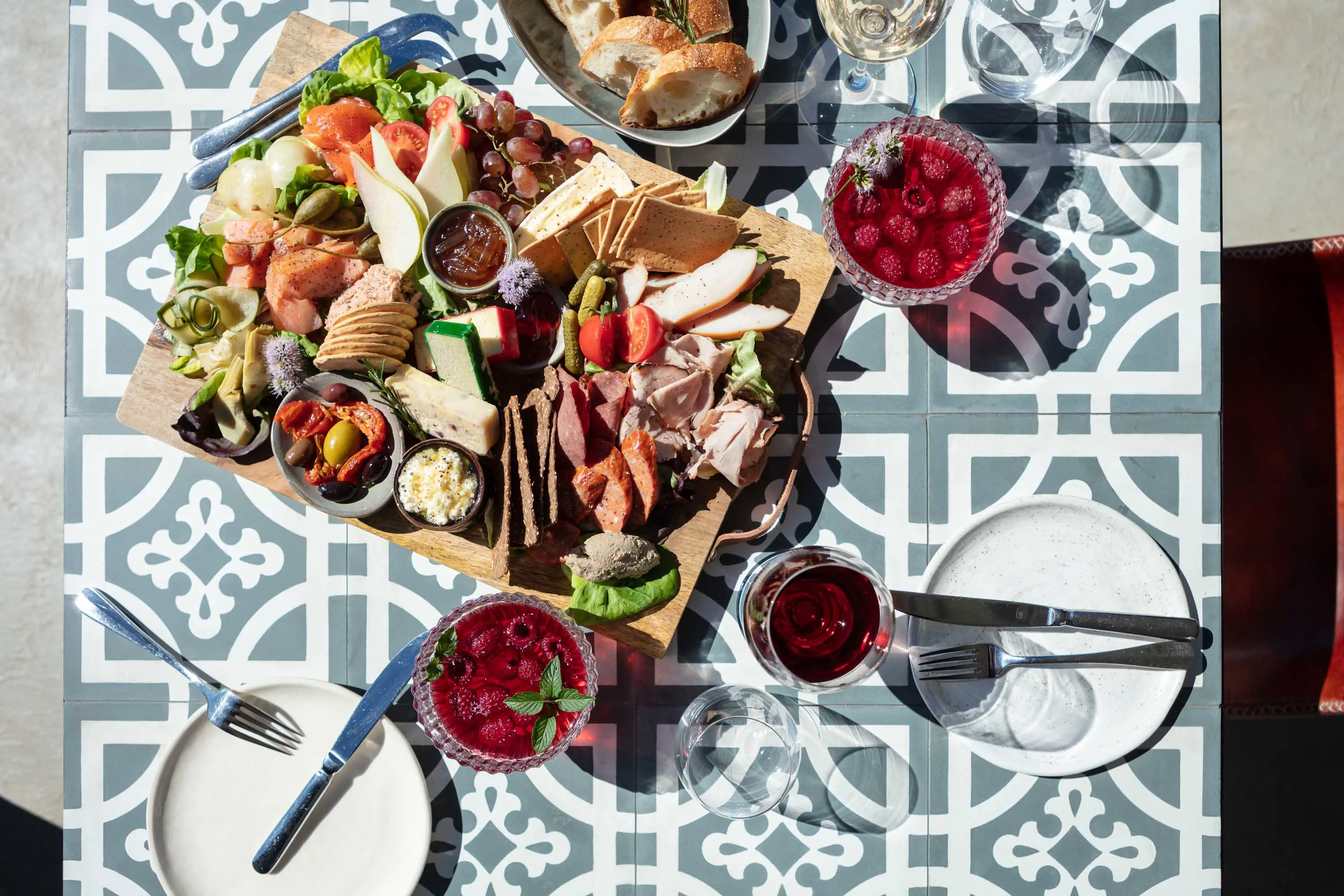 A mixed platter of cheese, dips, meats and fruits with dishes, cutlery and glasses filled with a raspberry dessert.