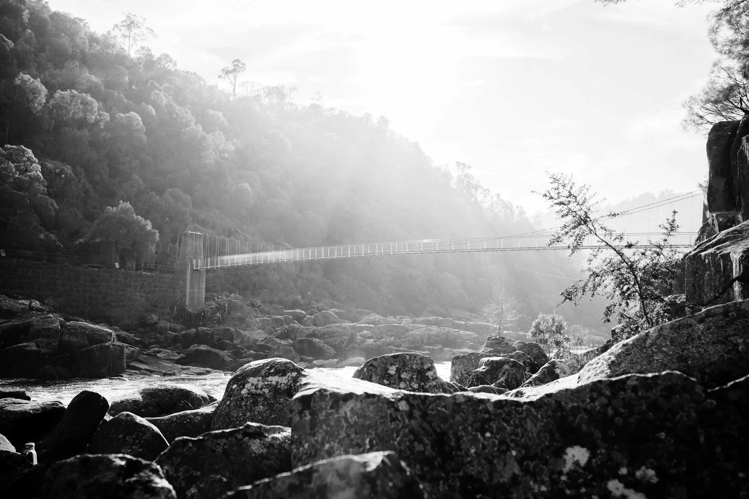 A long, thin bridge extends over a rocky river. Sunlight streams through clouds overhead.