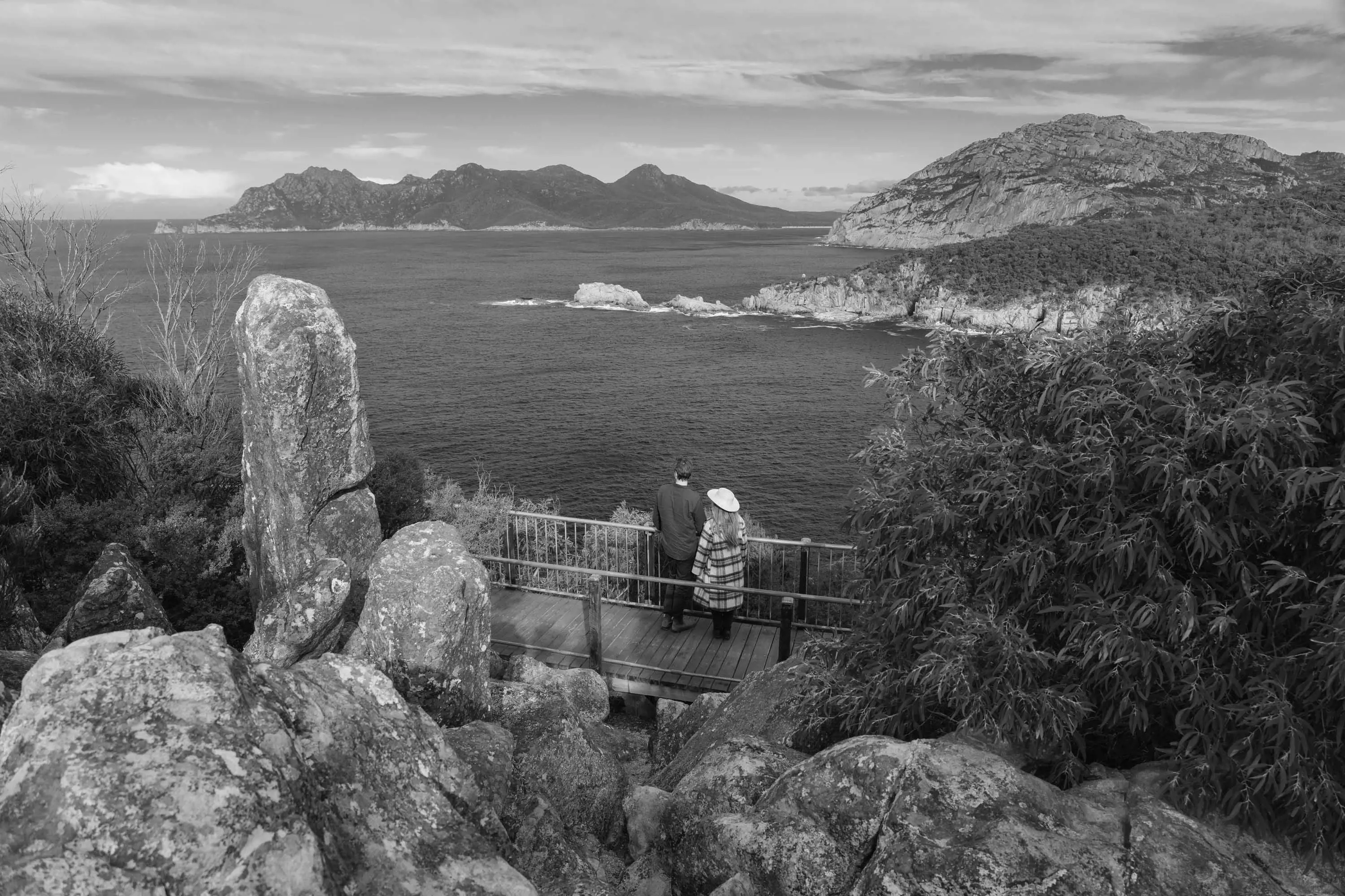 A young couple stand on the boardwalks on the edge of a coastal cliff looking out over water to a point on the horizon.