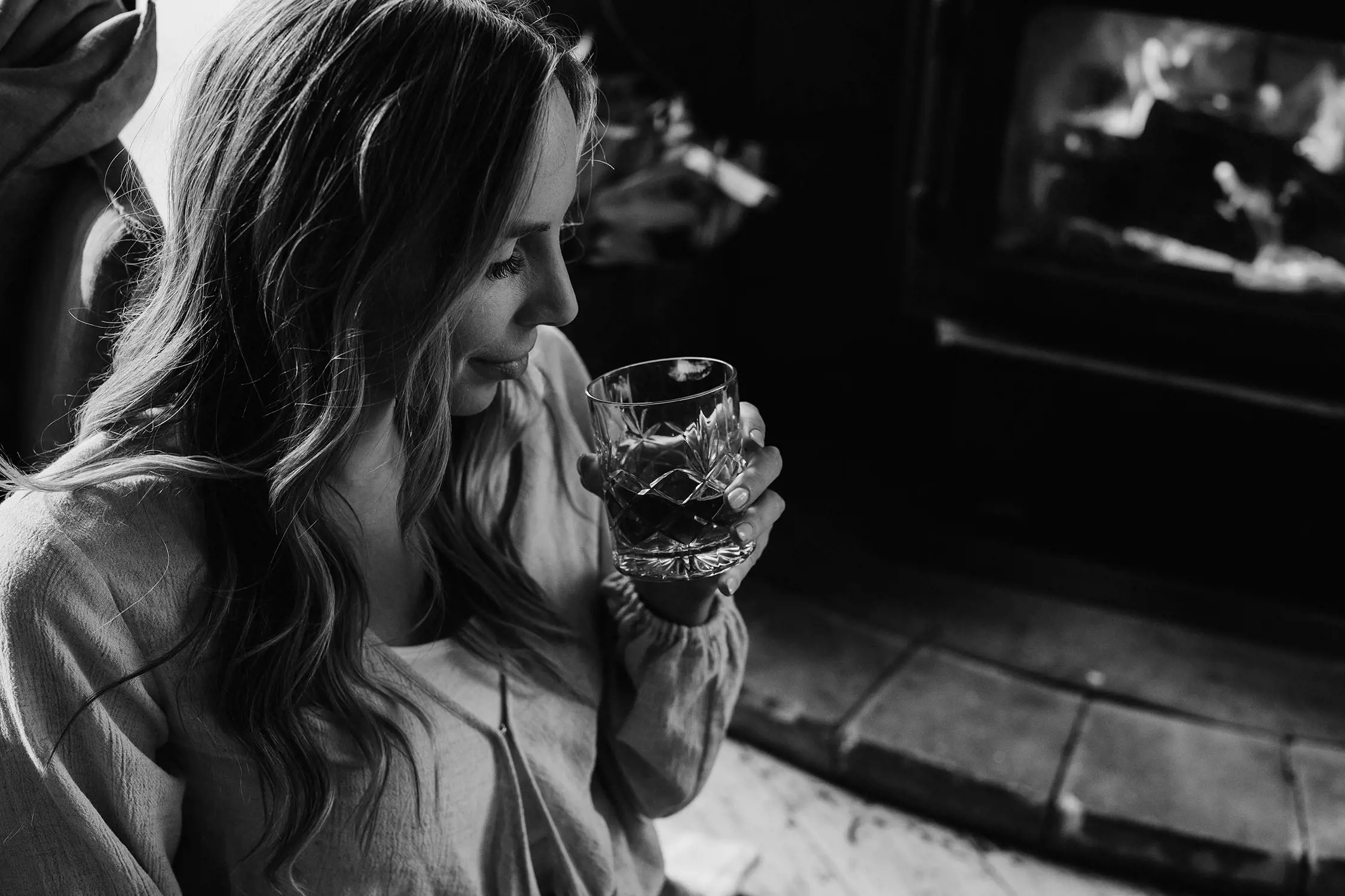 A woman holds a cut crystal whisky glass, relaxing and reclining by a roaring wood fireplace.