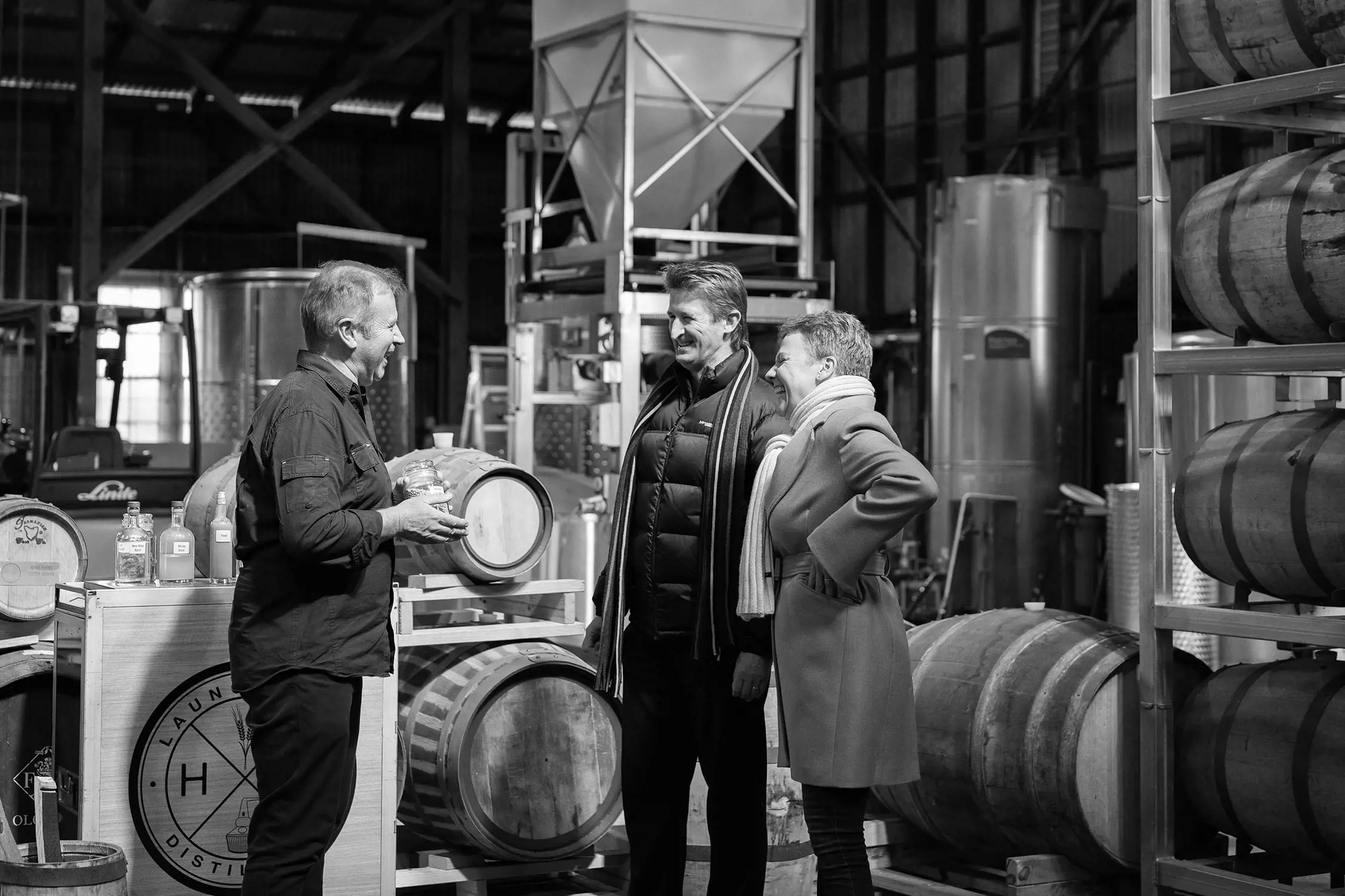 Inside a warehouse-style building, surrounded by whisky brewing barrels and equipment, a man talks to two guests.