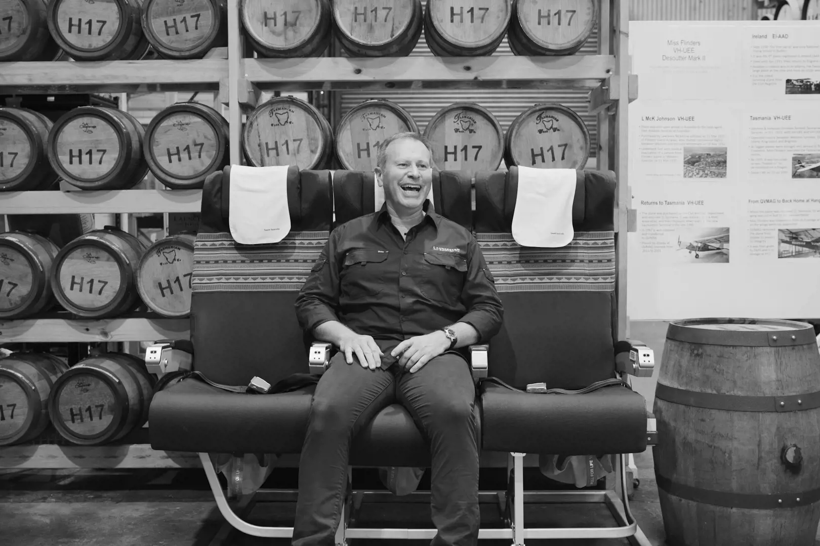 A man sits in the middle seat of a row of three airplane seats, laughing. The seats are placed in a warehouse in front of rows of whisky barrels on shelves.