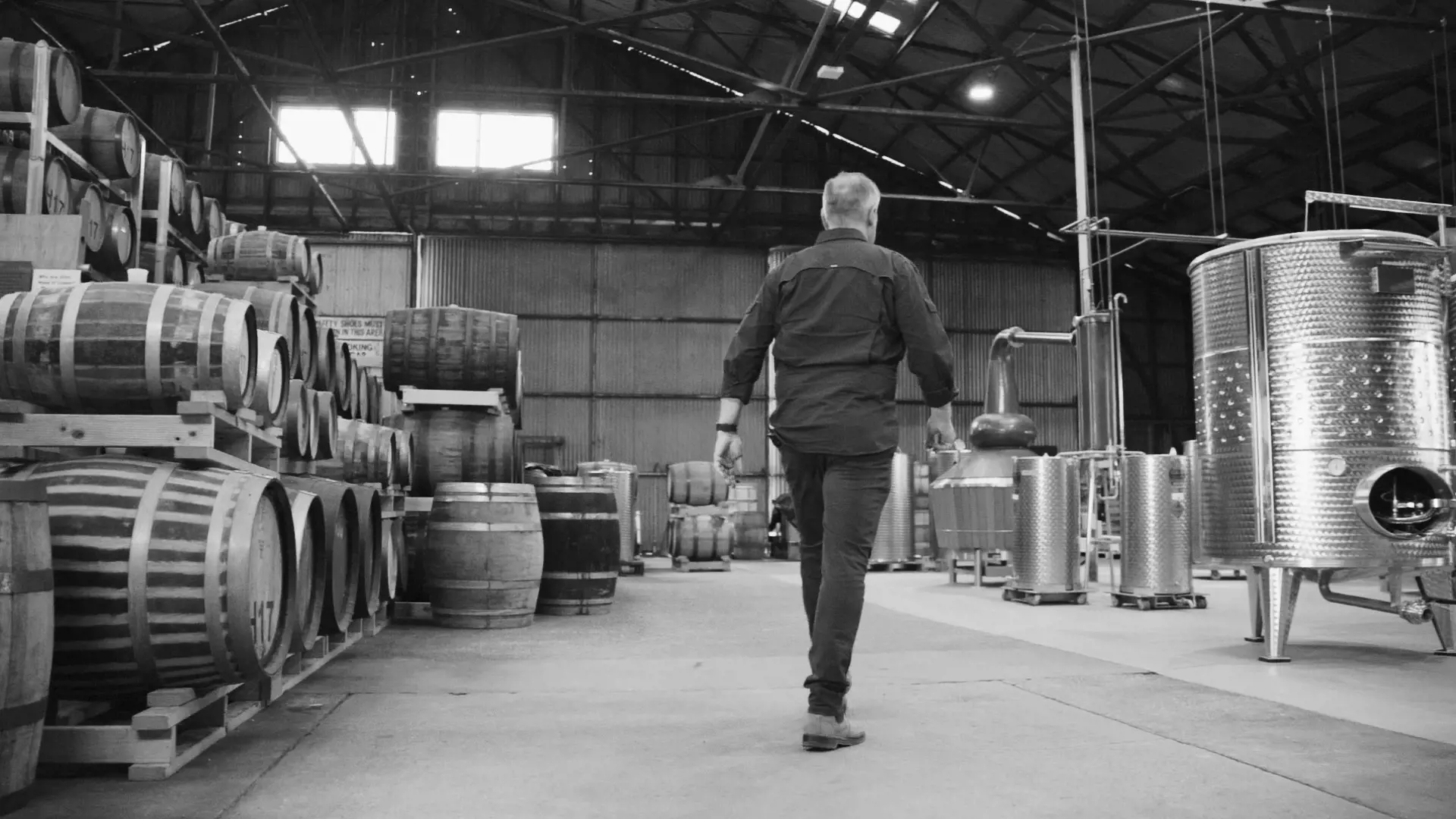 A man walks away from the camera into a warehouse space, filled with whisky barrels on shelving and distilling equipment.