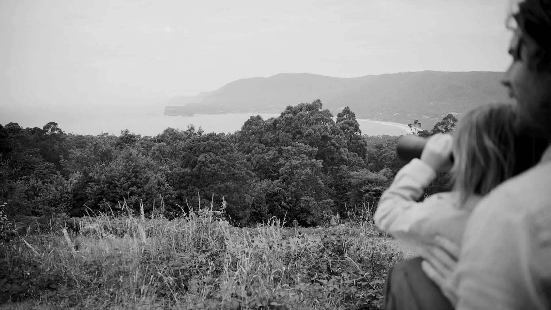 A child and an adult, both looking through binoculars, are situated outdoors surrounded by trees and a view of the coastline in the background.