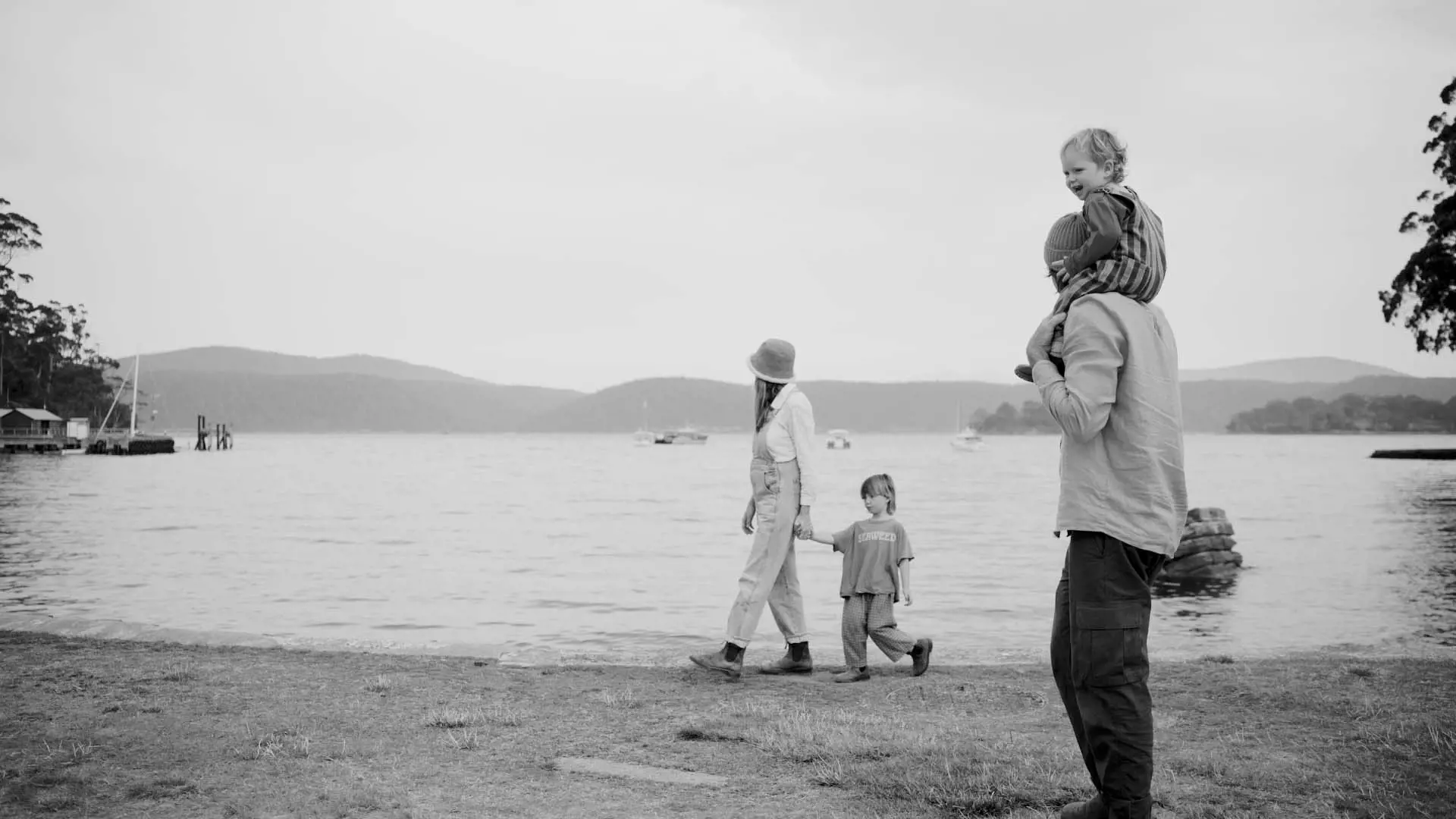 A family walks along the shore with one adult carrying a child on their shoulders. The background features water and grassy areas against a misty landscape.