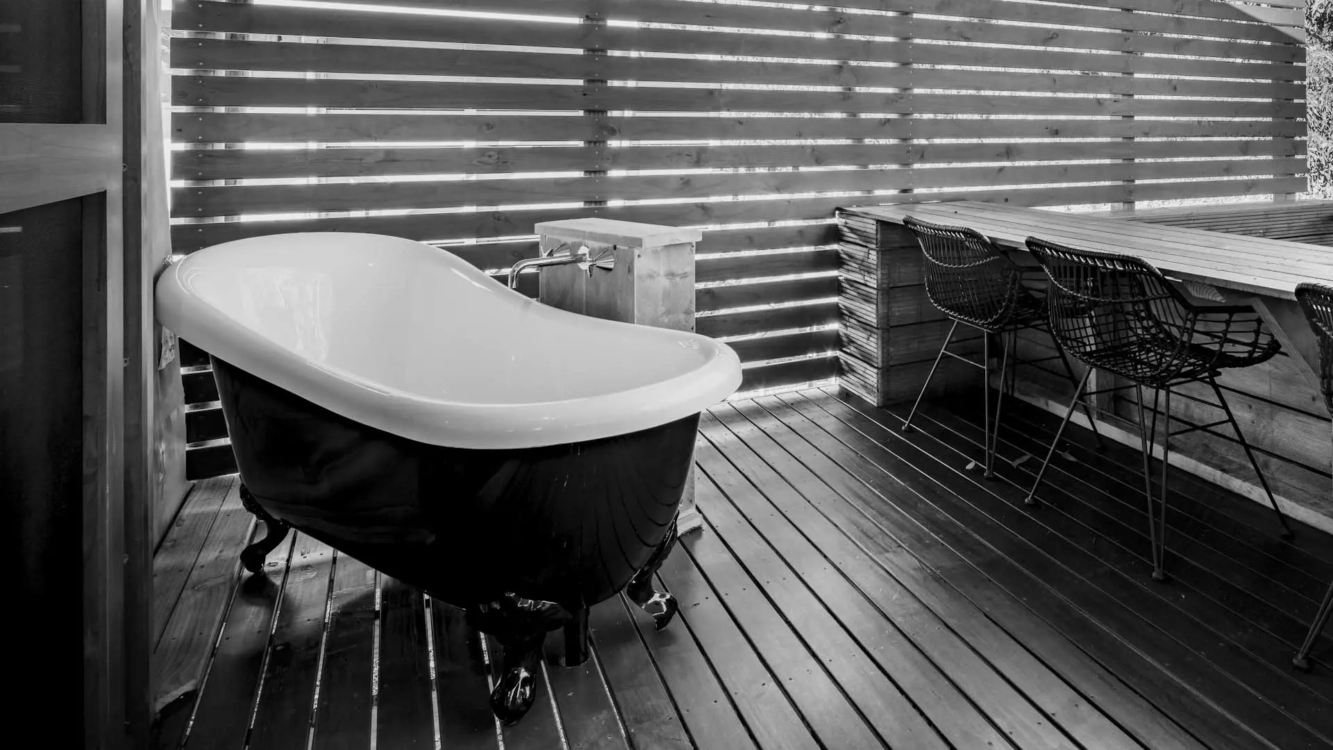A clawfoot bathtub is placed in a rustic outdoor setting, with shaded wooden slats allowing natural light to filter through. Nearby, there is a table and chairs.