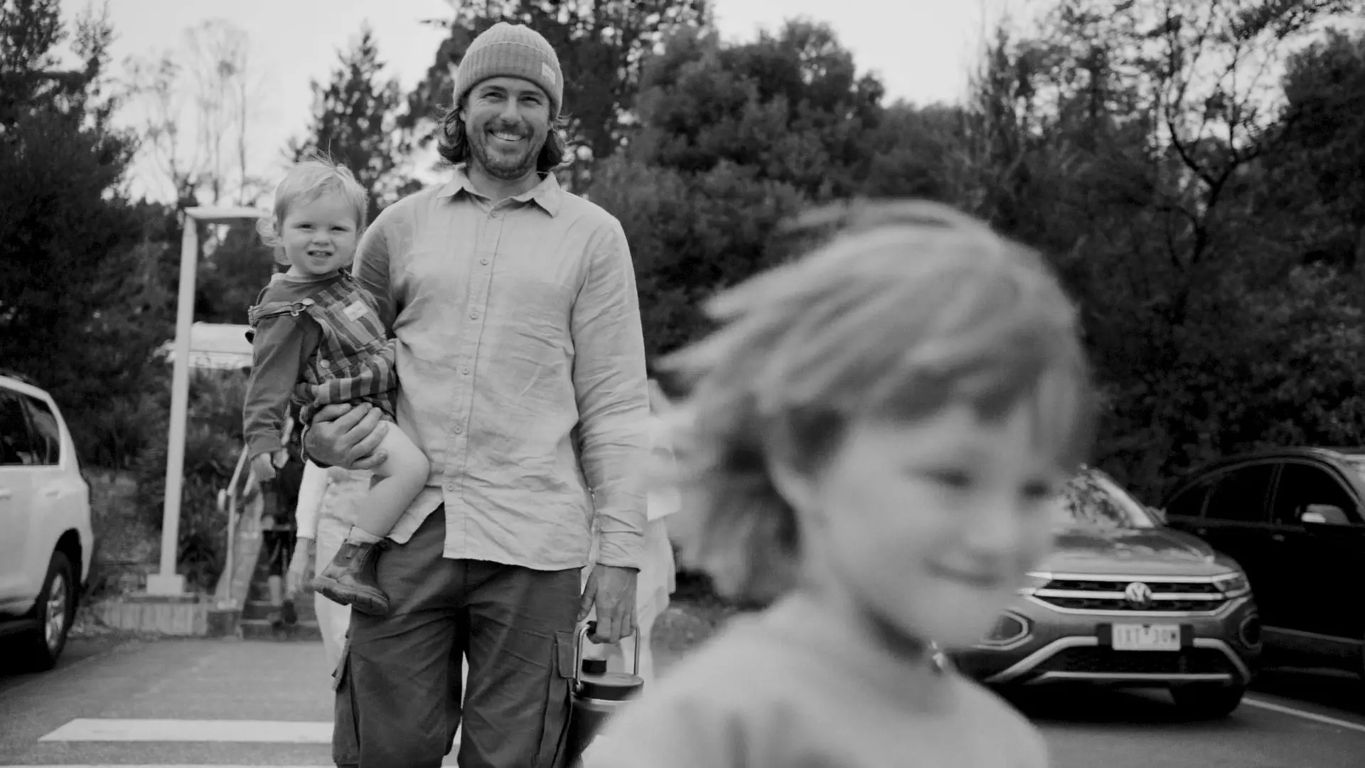 In a carpark, a father holding his toddler on his hip smiles looking at his other child running toward the camera.