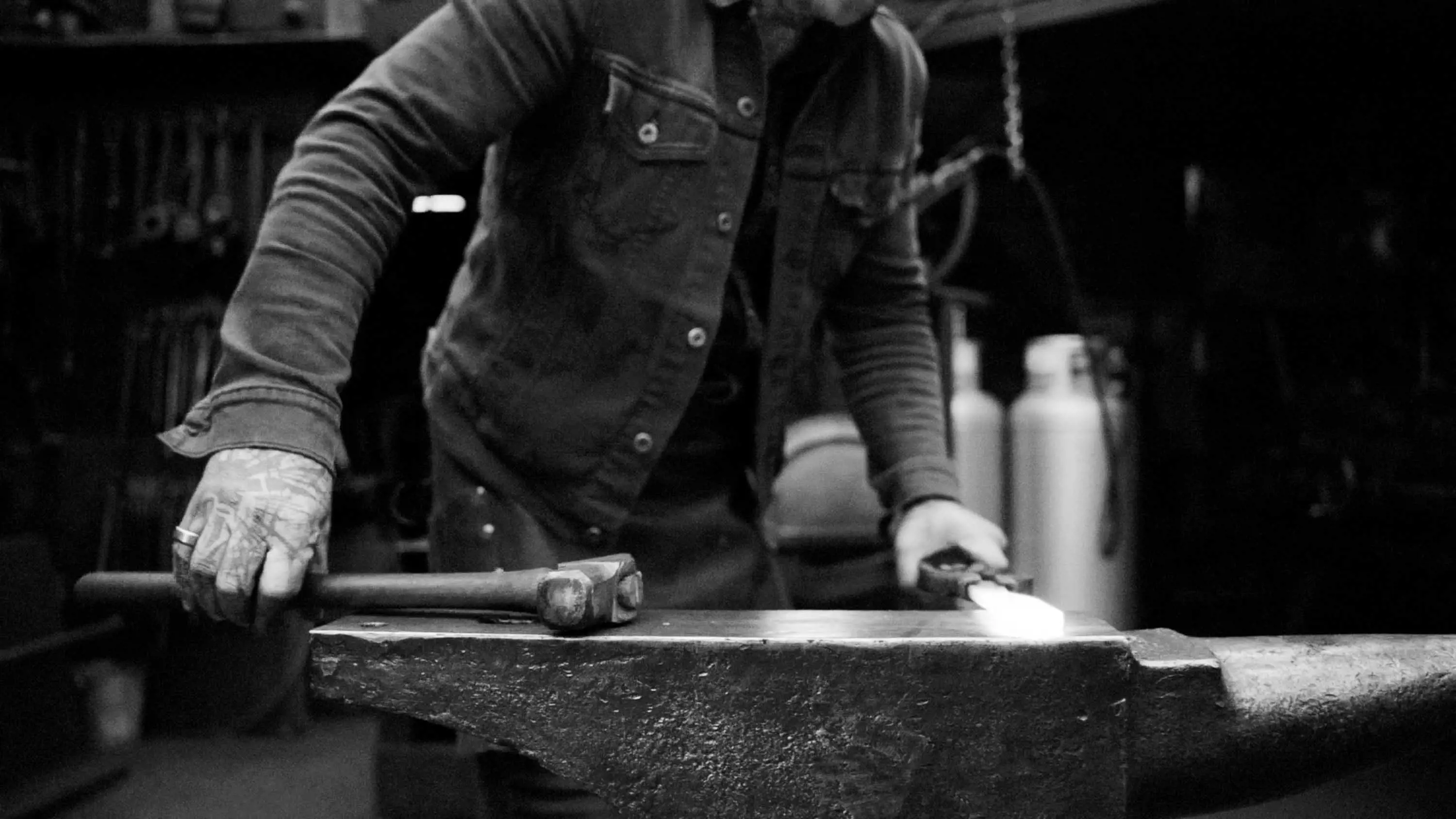 A man wearing denim and a leather blacksmiths apron picks up a hammer next to a piece of white-hot steel held in a pair or pichers.