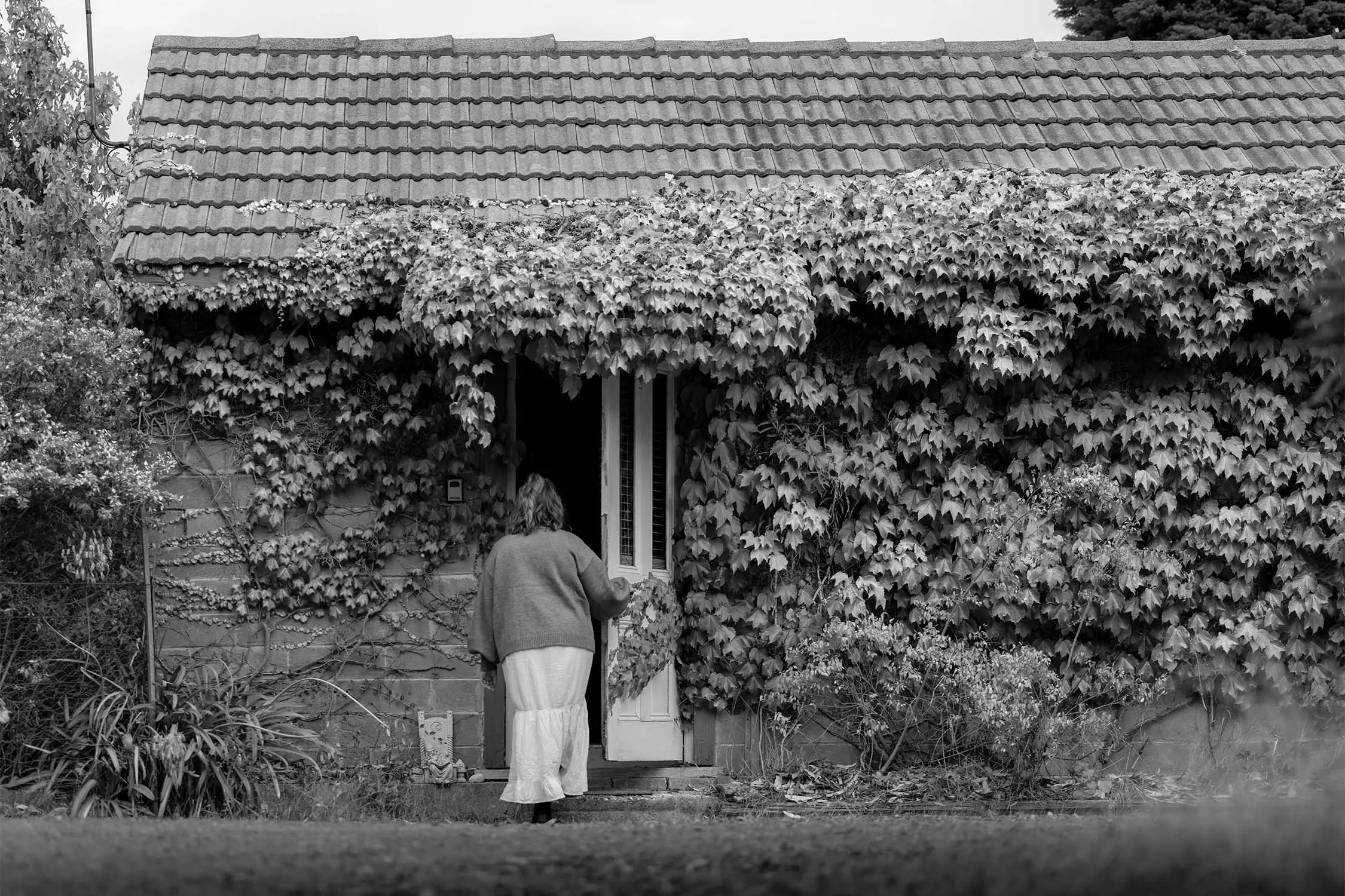 A woman in a sweater walks toward a house covered in ivy, with a tiled roof. The scene is set outdoors and depicted in black and white.