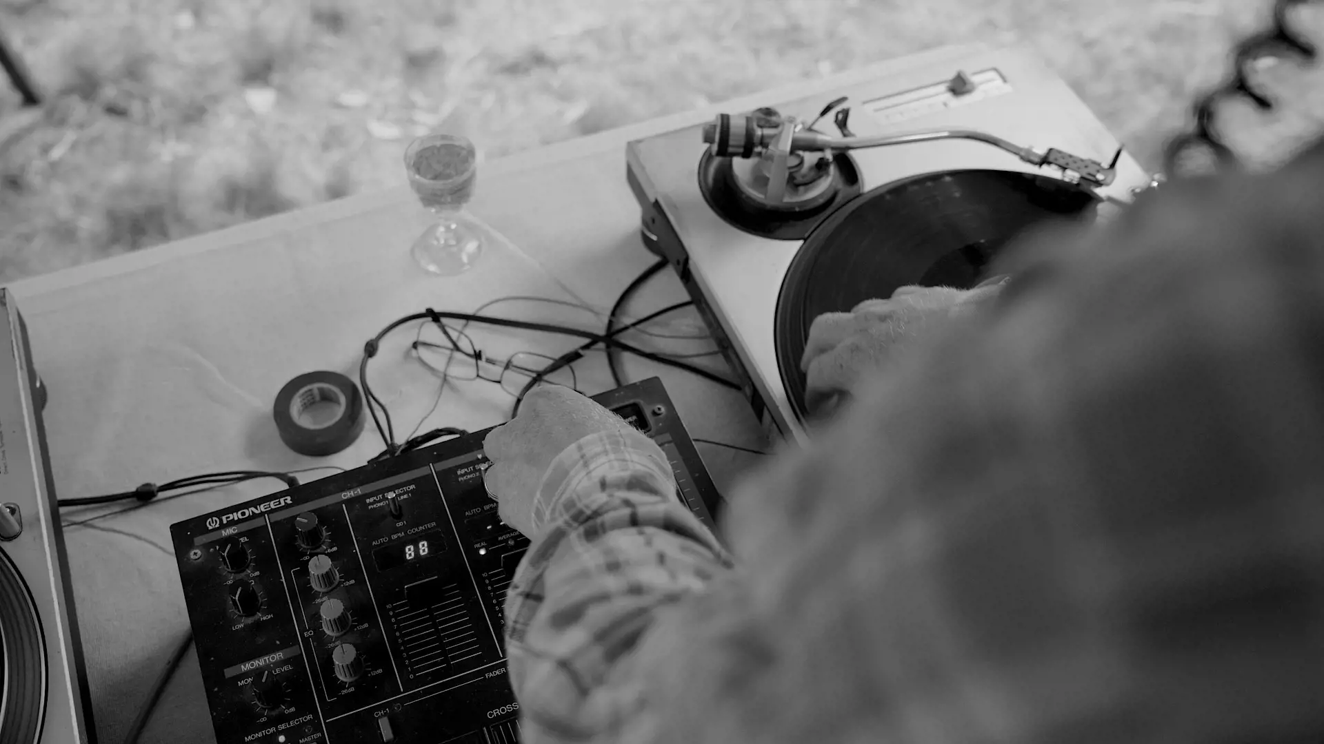 Looking down over the shoulder of a person wearing a plaid shirt, who is spinning a vinyl record with one hand and turning knobs on a mixing desk with another.