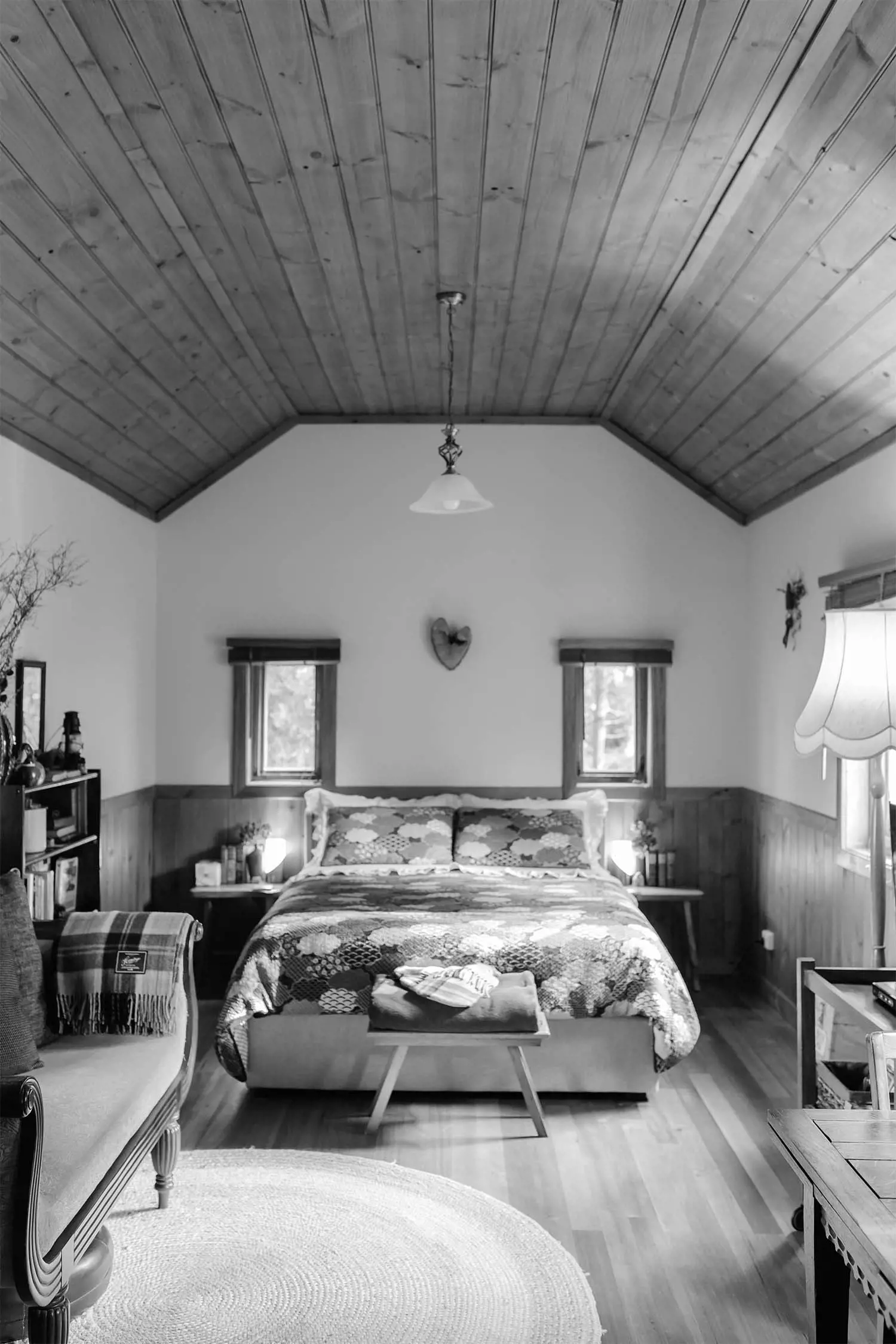 An interior view of a cozy bedroom featuring a bed with patterned bedding, wooden accents, and soft lighting. The room has a warm atmosphere, captured in black and white.