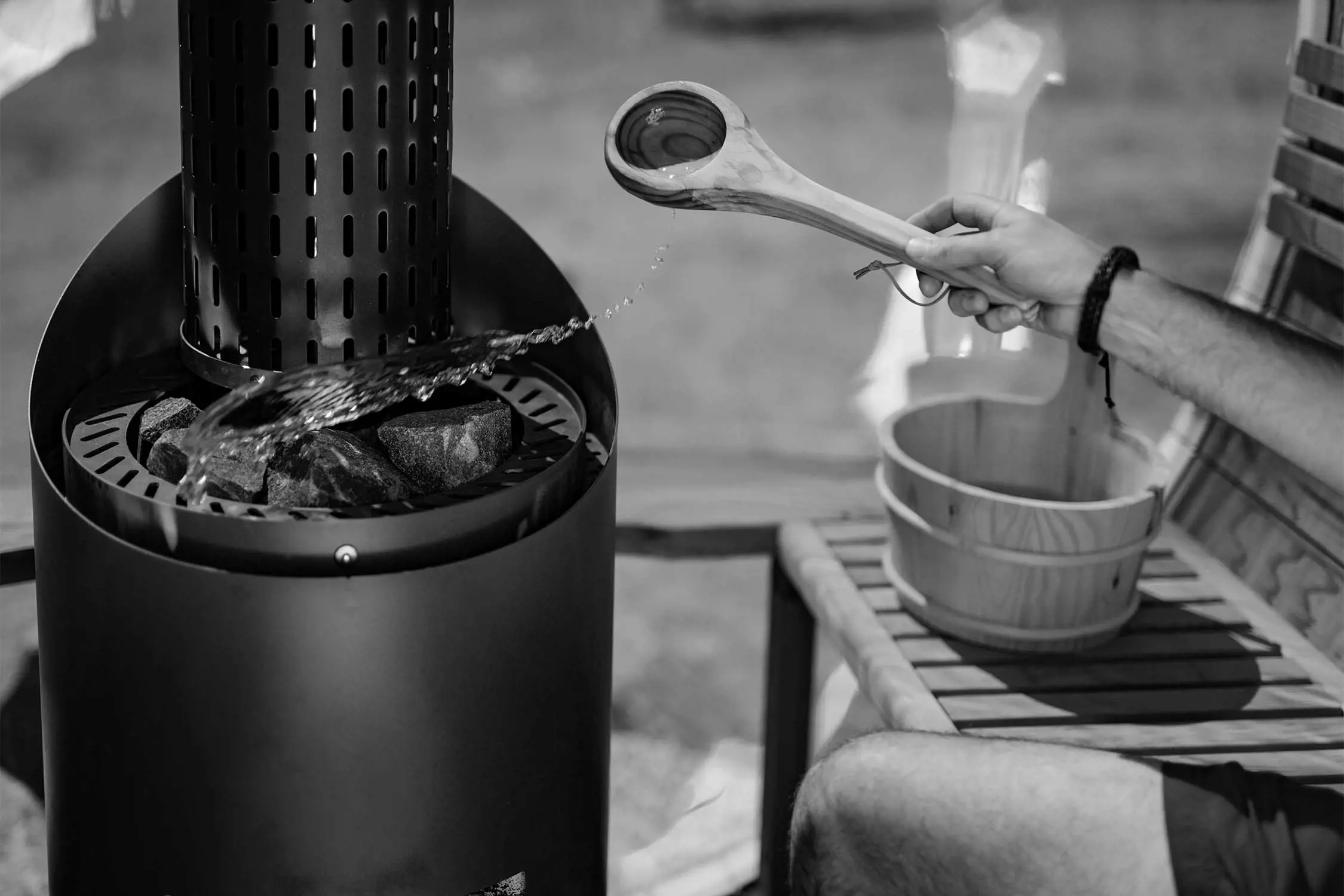 A person sits on a wooden bench beside a small furnace, adding a splash of water over the hot coals using a hand-carved wooden spoon.