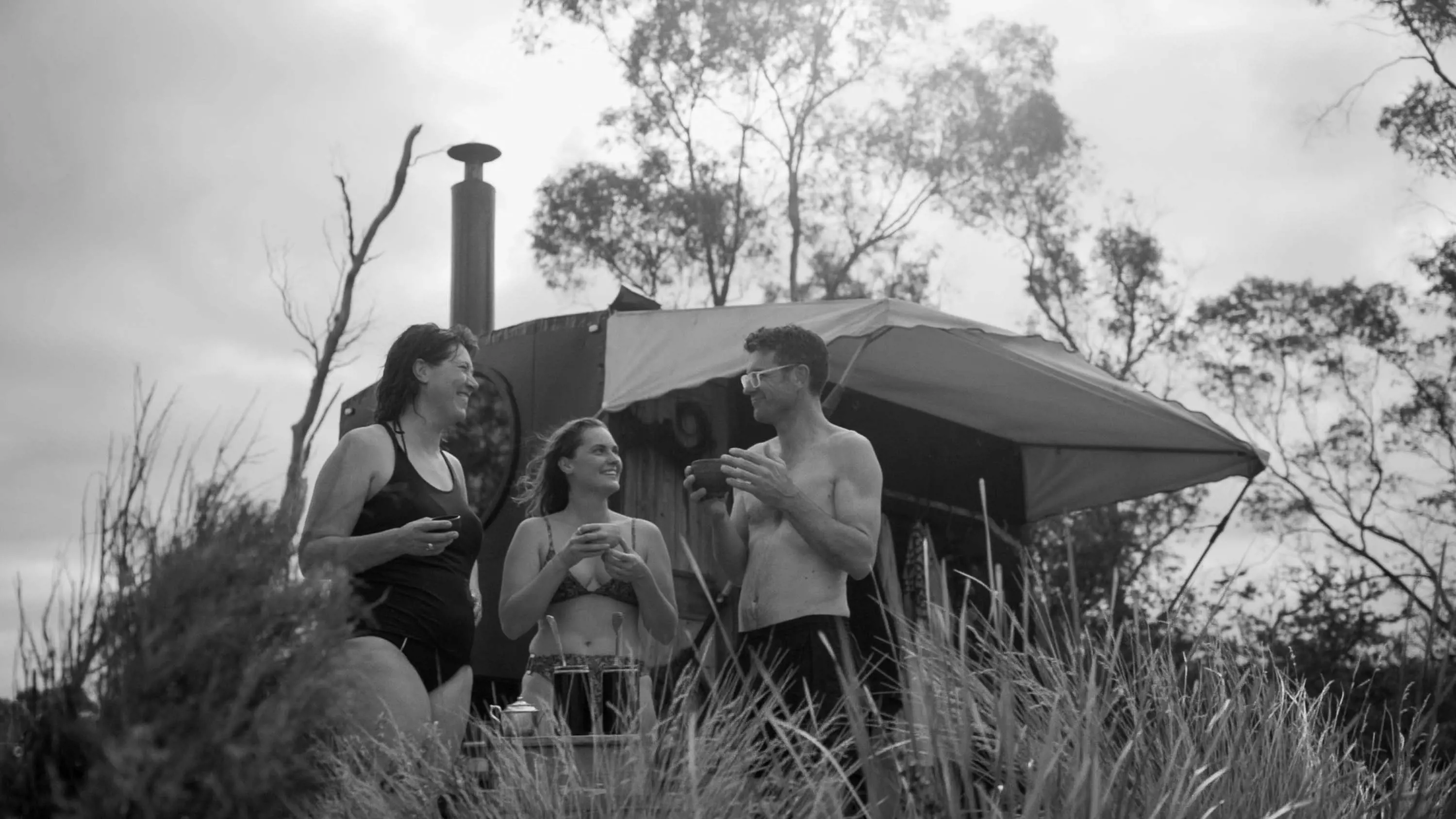 3 people stand in front of a portable sauna, parked beneath the shade of trees.
