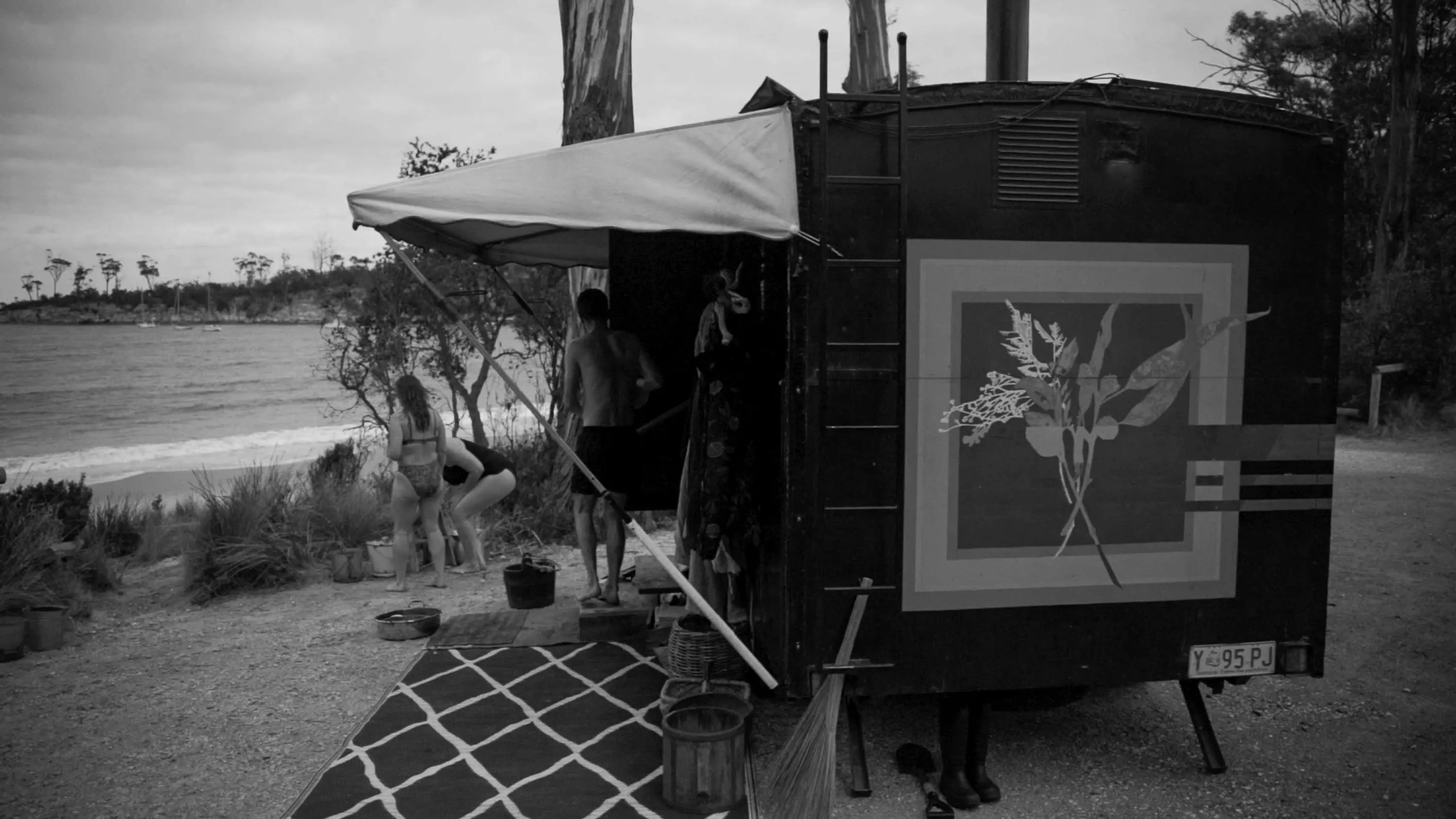 A portable sauna built on a trailer with a canopy and tall chimney is parked near a beach.