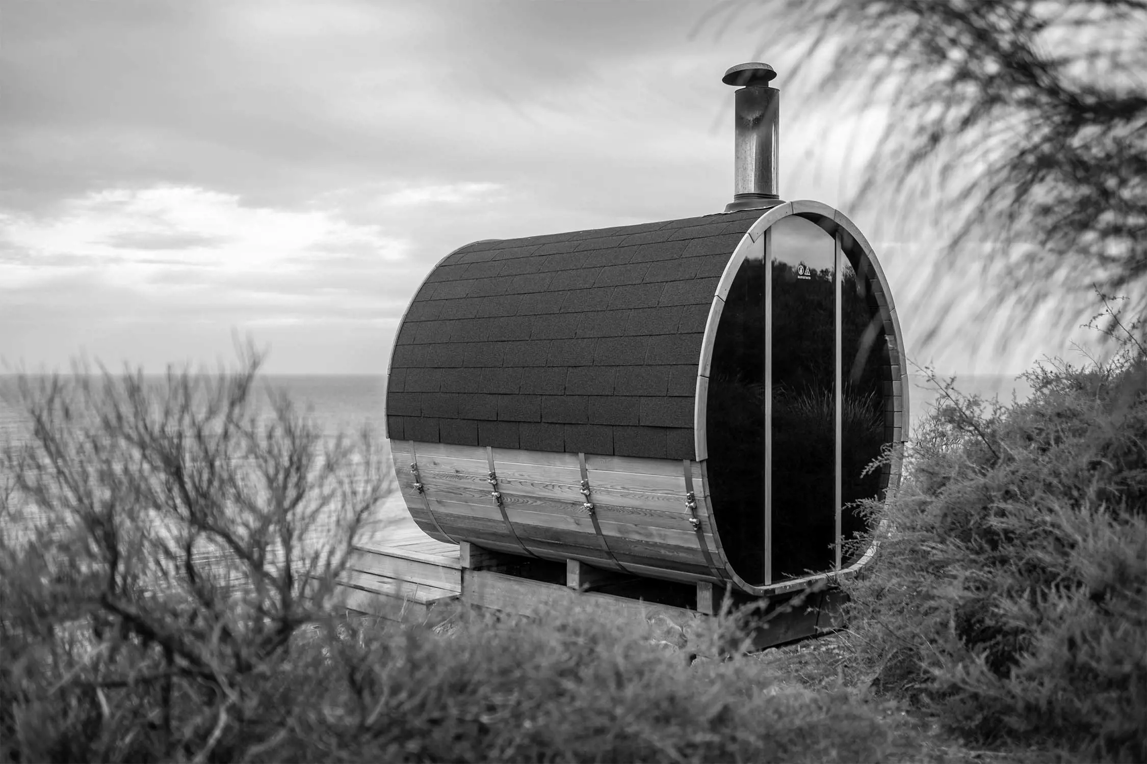 A modern barrel-shaped structure with a metal chimney, surrounded by shrubs, overlooking a body of water under a cloudy sky. The image is in black and white