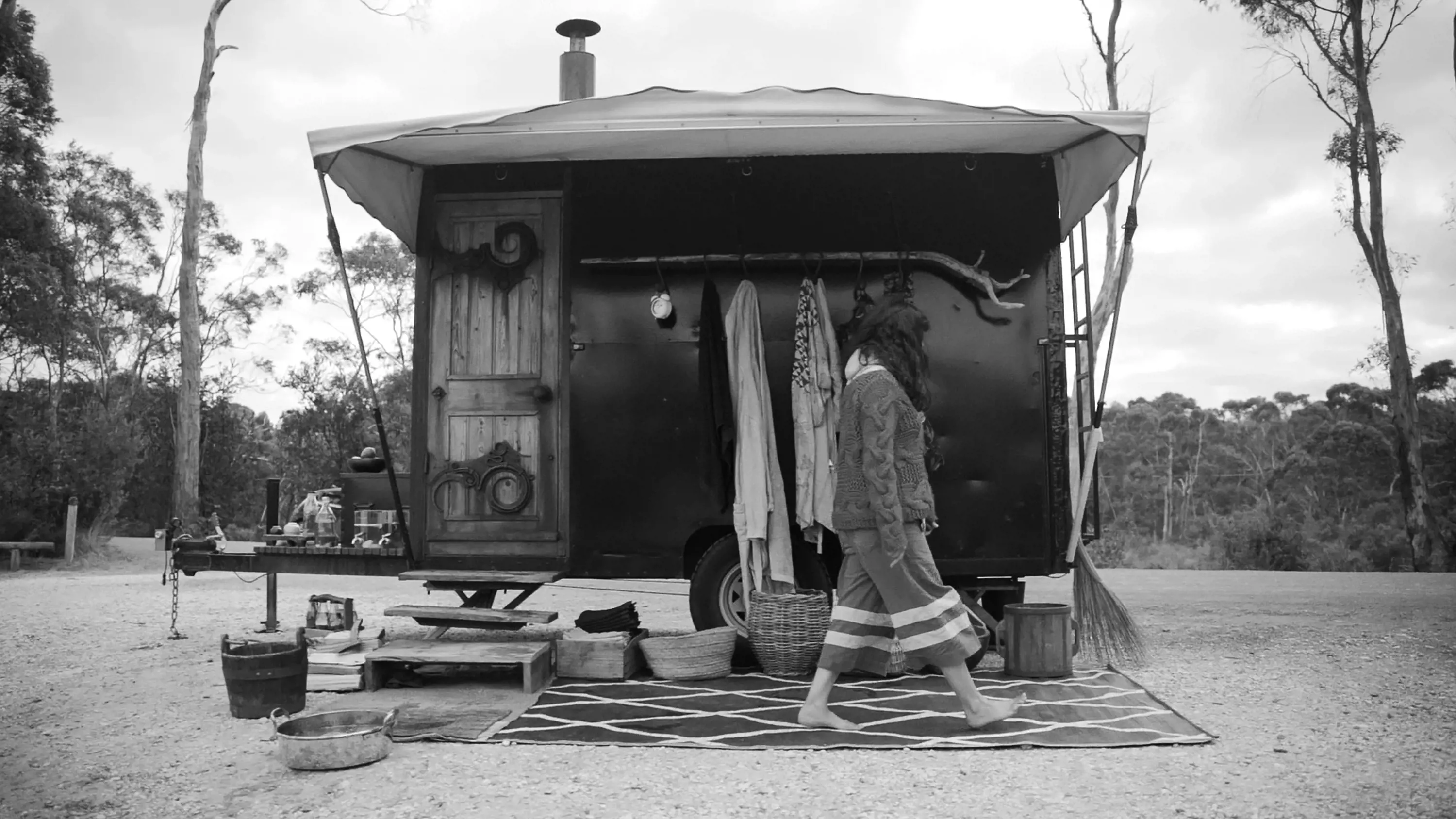 A woman walks by a portable sauna on a trailer, set under trees at a beach.