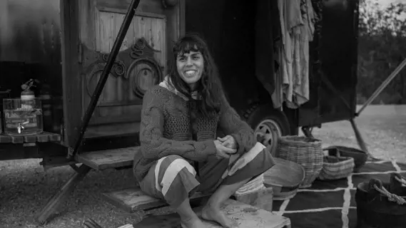 A woman wearing a loose-nit woollen sweater and half-cut pants sits on the steps of a portable sauna built on a trailer.