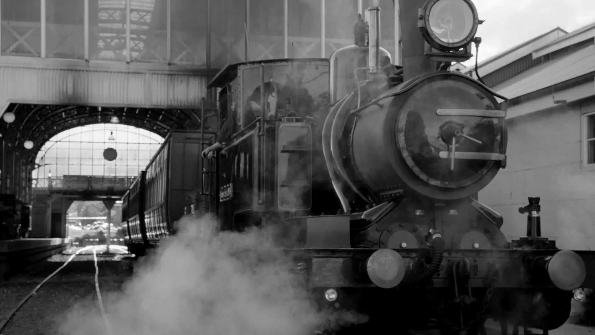 A large steam locomotive with passenger carriages exits the station with a puff of steam.