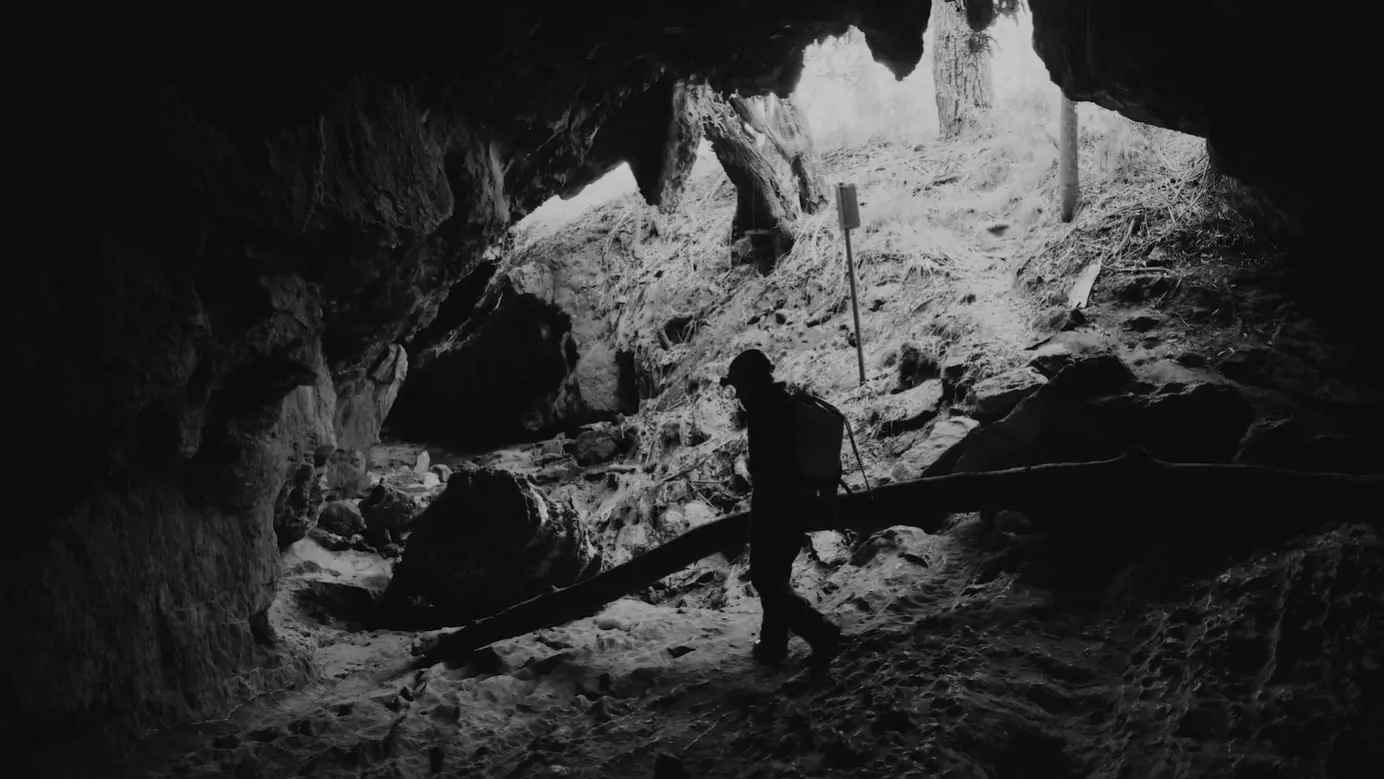 A figure walks in the base of a rocky cave system illuminated by the light of an overhead opening.