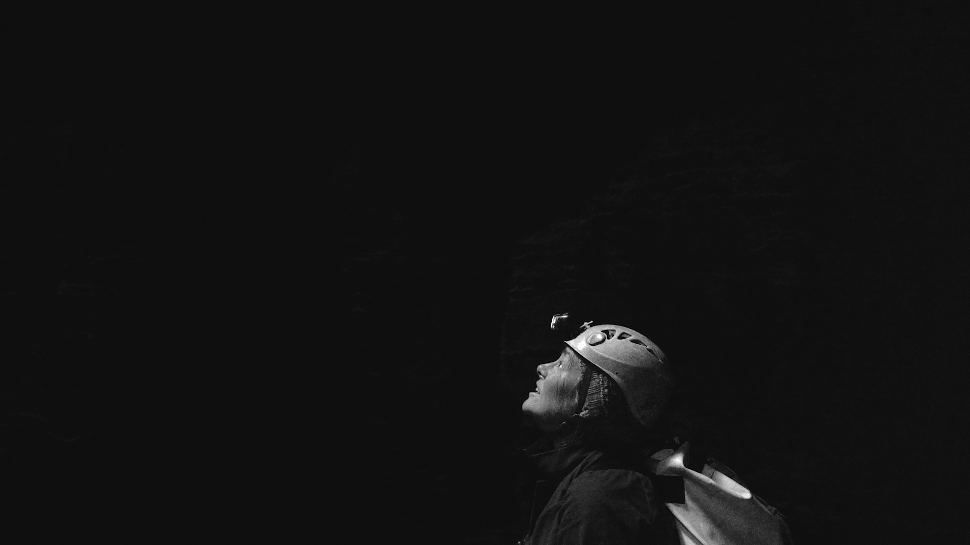 A woman with a hard hat and light looks upwards in the pitch darkness of a cave.