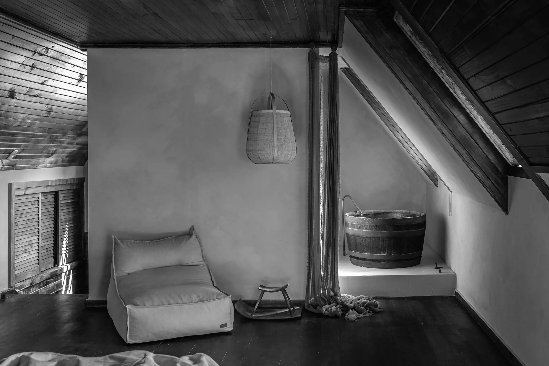 A cozy room with wooden ceilings and floors. There is a large bean bag chair and a wicker basket hanging from the ceiling. In the corner is a rustic wooden bathtub on a raised platform with curtains partially drawn.