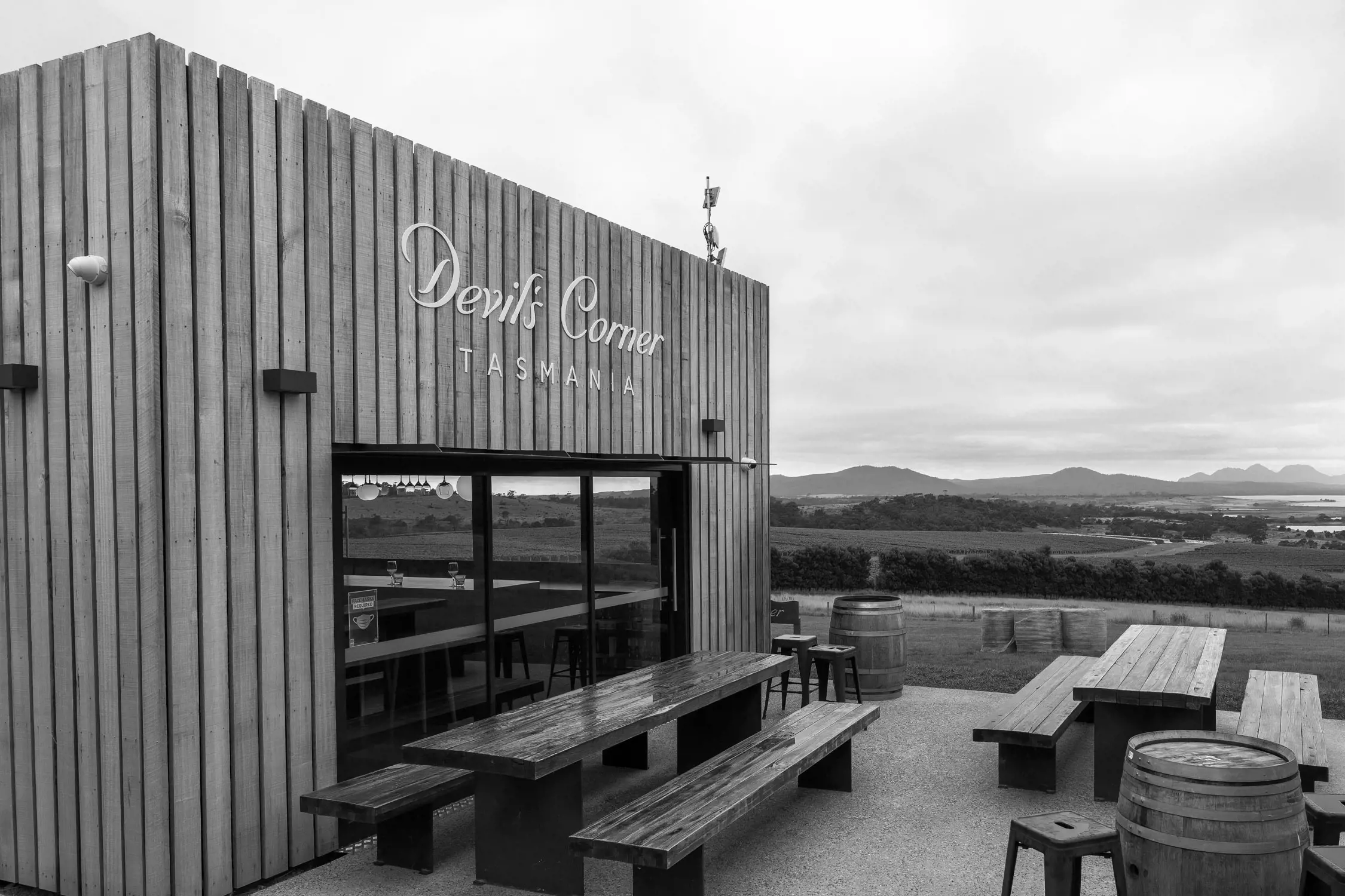 A stylish wooden winery building with a large sign reading 'Devil’s Corner Tasmania.' The structure features modern architecture with clean lines, large windows, and a spacious outdoor seating area with polished wooden picnic tables and barrels. Beyond the building, rolling vineyards and distant hills stretch under a cloudy sky
