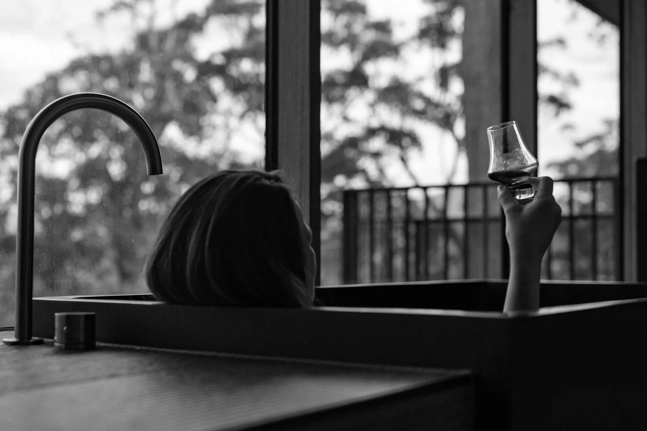 A person reclines in a bathtub near a large window, holding a glass of wine raised in the air. Outside, trees fill the serene view, evoking a peaceful connection to nature