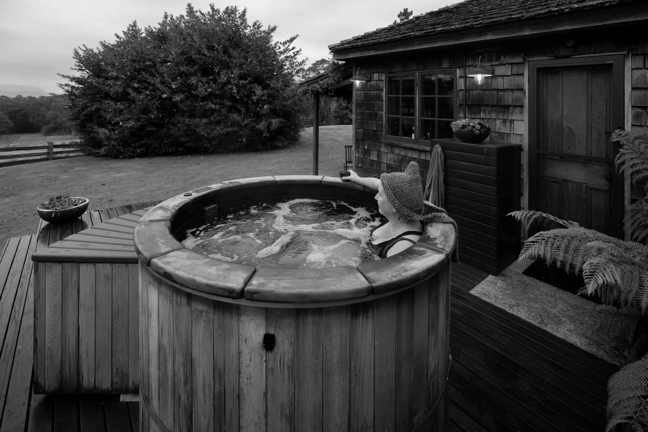 A serene scene showcasing a person relaxing in a warm wooden hot tub outdoors. The tub is surrounded by verdant fields and distant hills under a cloudy sky, creating a peaceful retreat-like atmosphere.