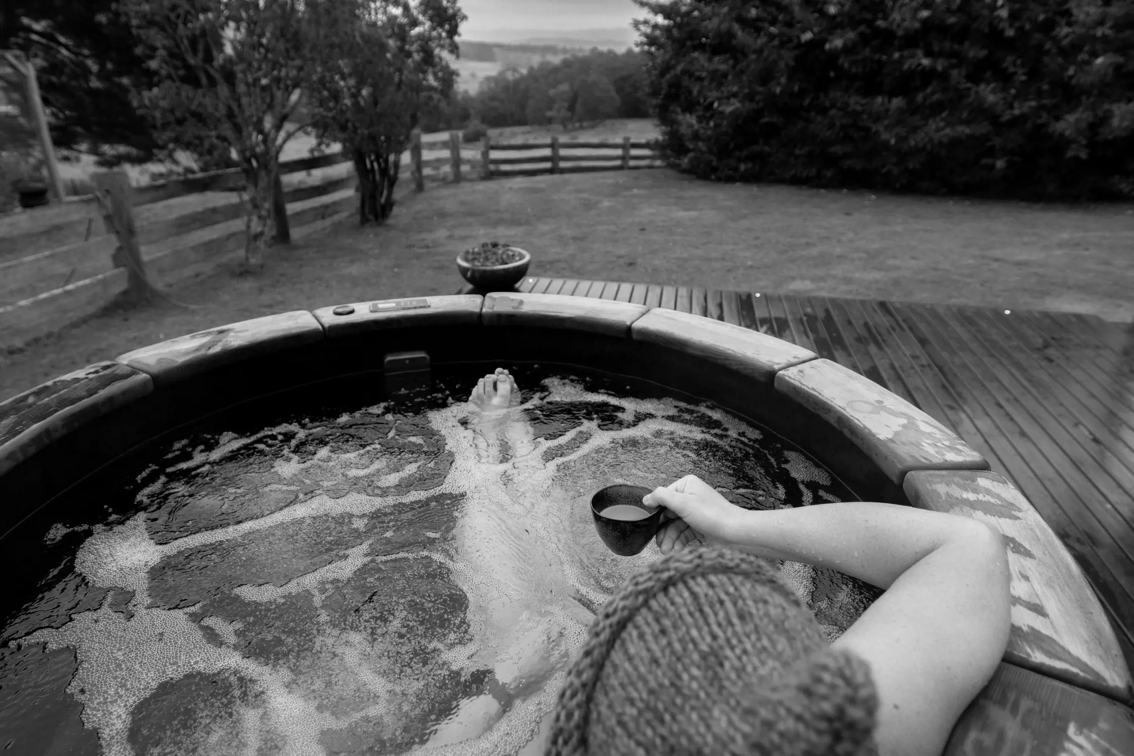 A wooden hot tub set outdoors in a tranquil countryside setting. A person is seen relaxing in the steaming water, gazing toward rolling hills and open fields. A wooden deck and a rustic building nearby add to the cozy atmosphere.