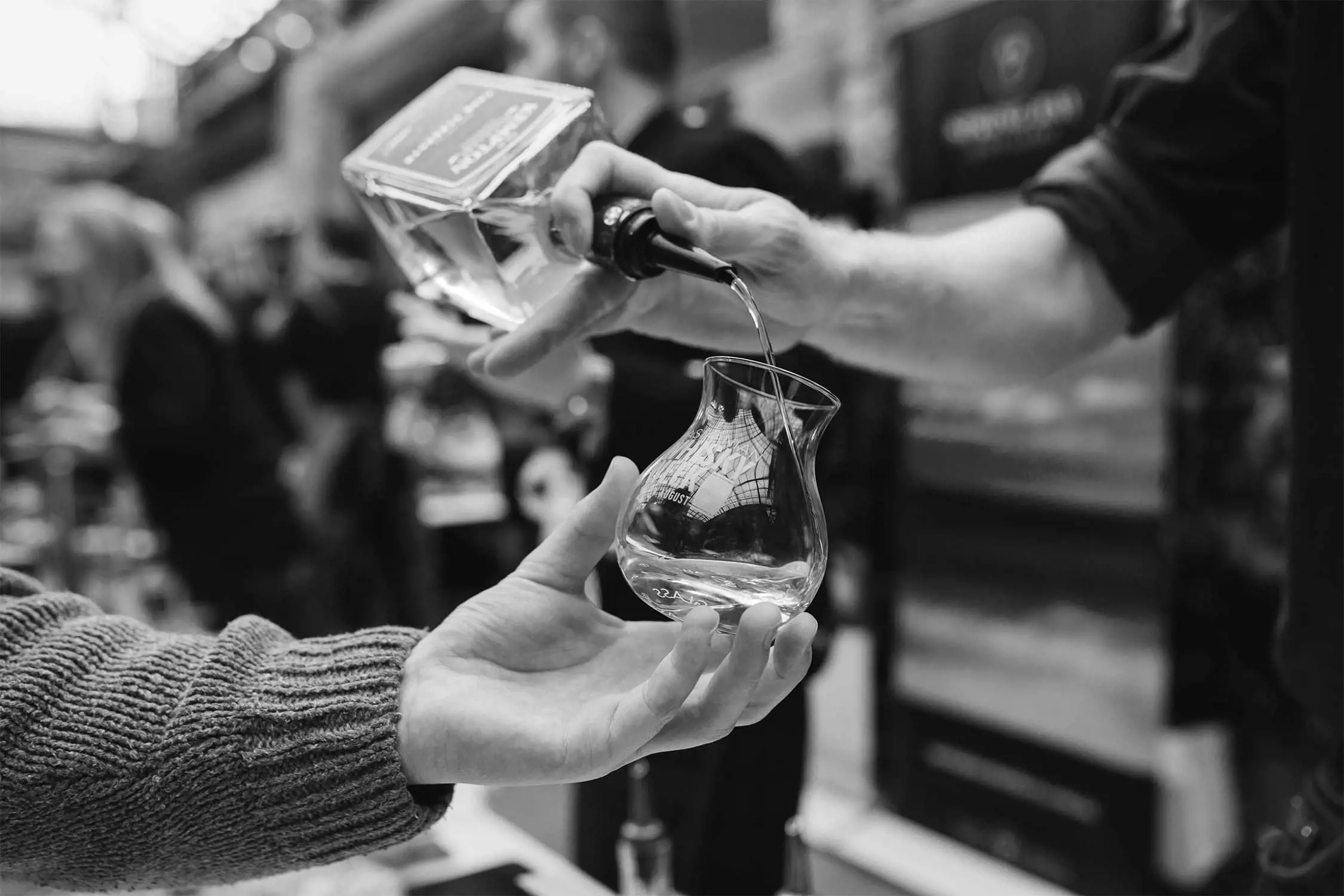 A close-up of a hand holding a curved glass vessel while another hand pours a clear liquid from a bottle into it. The scene is set in a busy environment, possibly at a tasting event, with blurred figures of people in the background. The image is in black and white