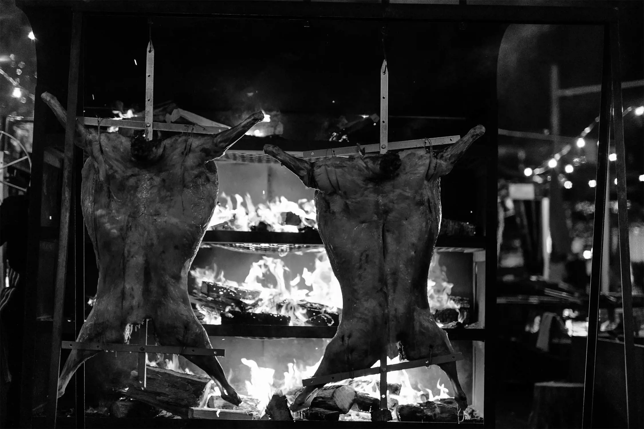 Two whole animals are being roasted over an open fire on a grill, surrounded by flames and smoke