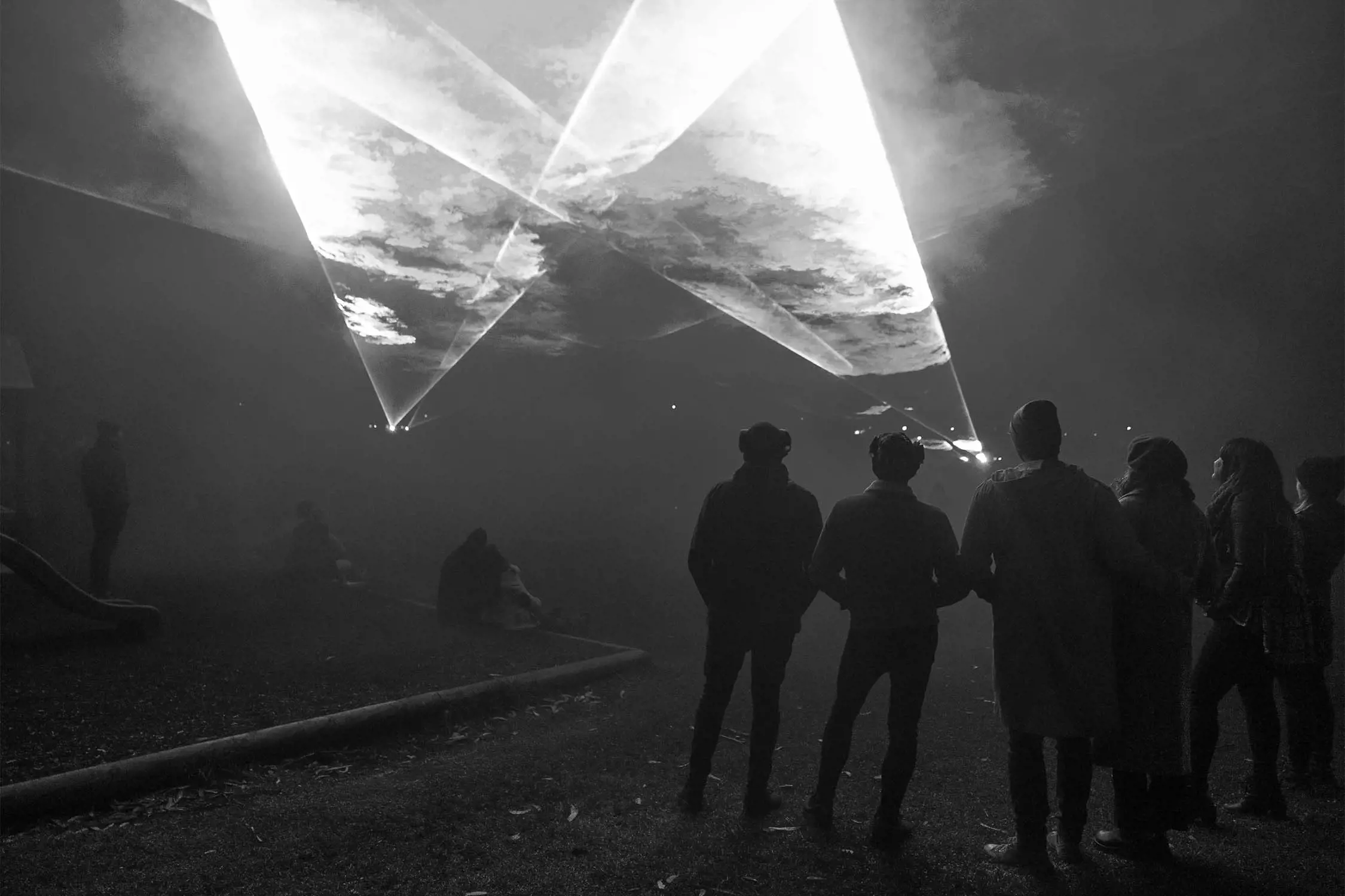 A group of people standing in a dark outdoor setting, watching laser beams crossing the sky, creating dramatic light patterns.