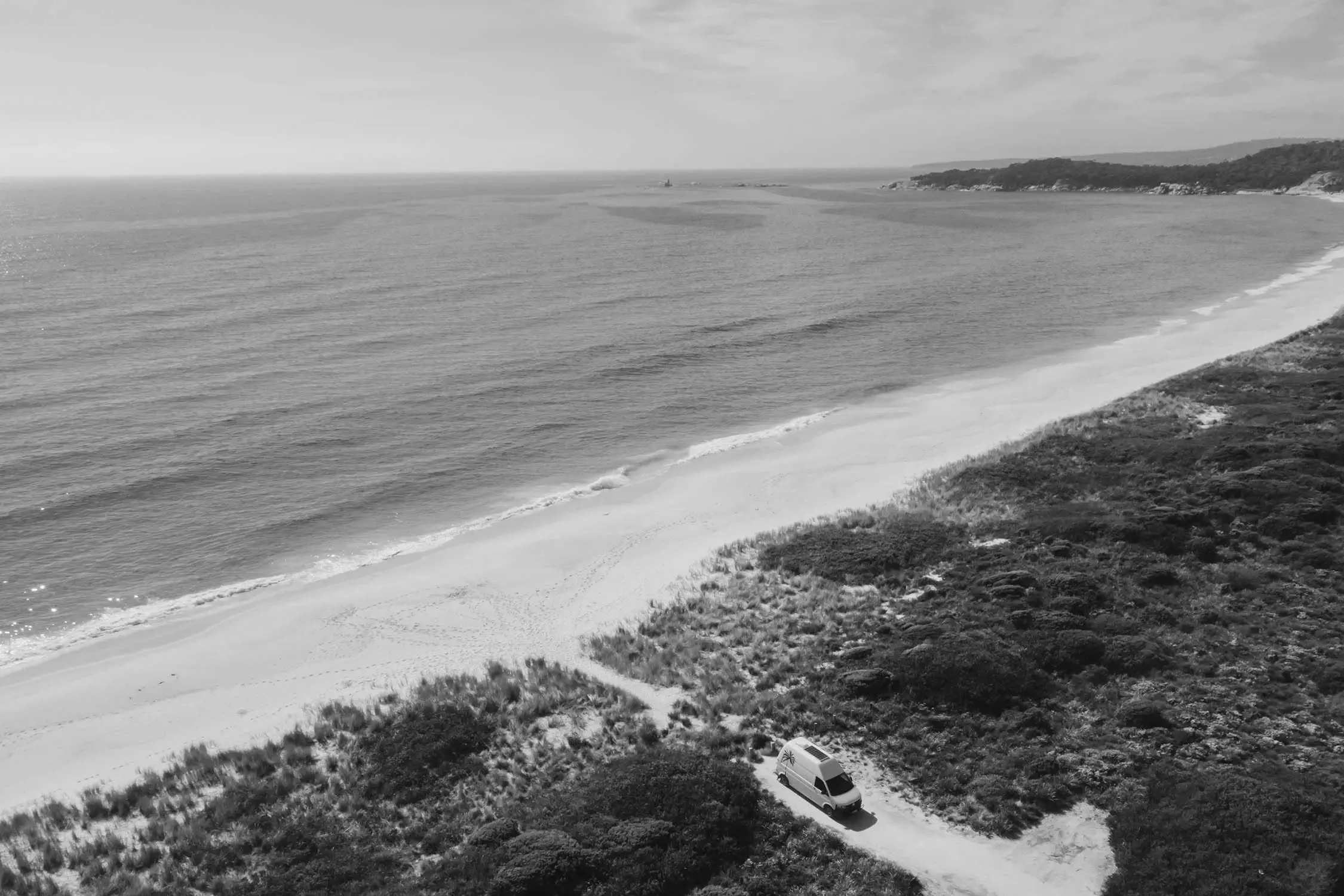 A serene black and white coastal scene featuring a white van parked near a sandy beach, with gentle waves lapping at the shore and hills in the background