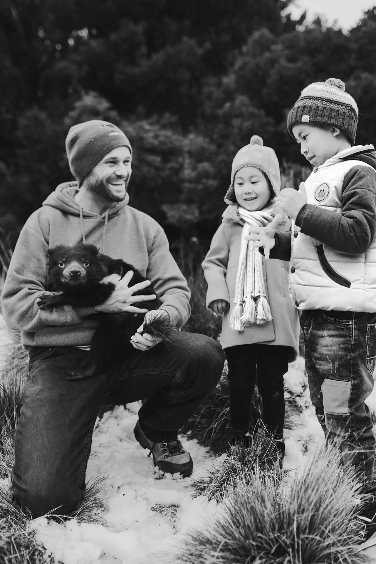 A man kneels with a black Tasmanian devil in his arms, while two children smile beside him.