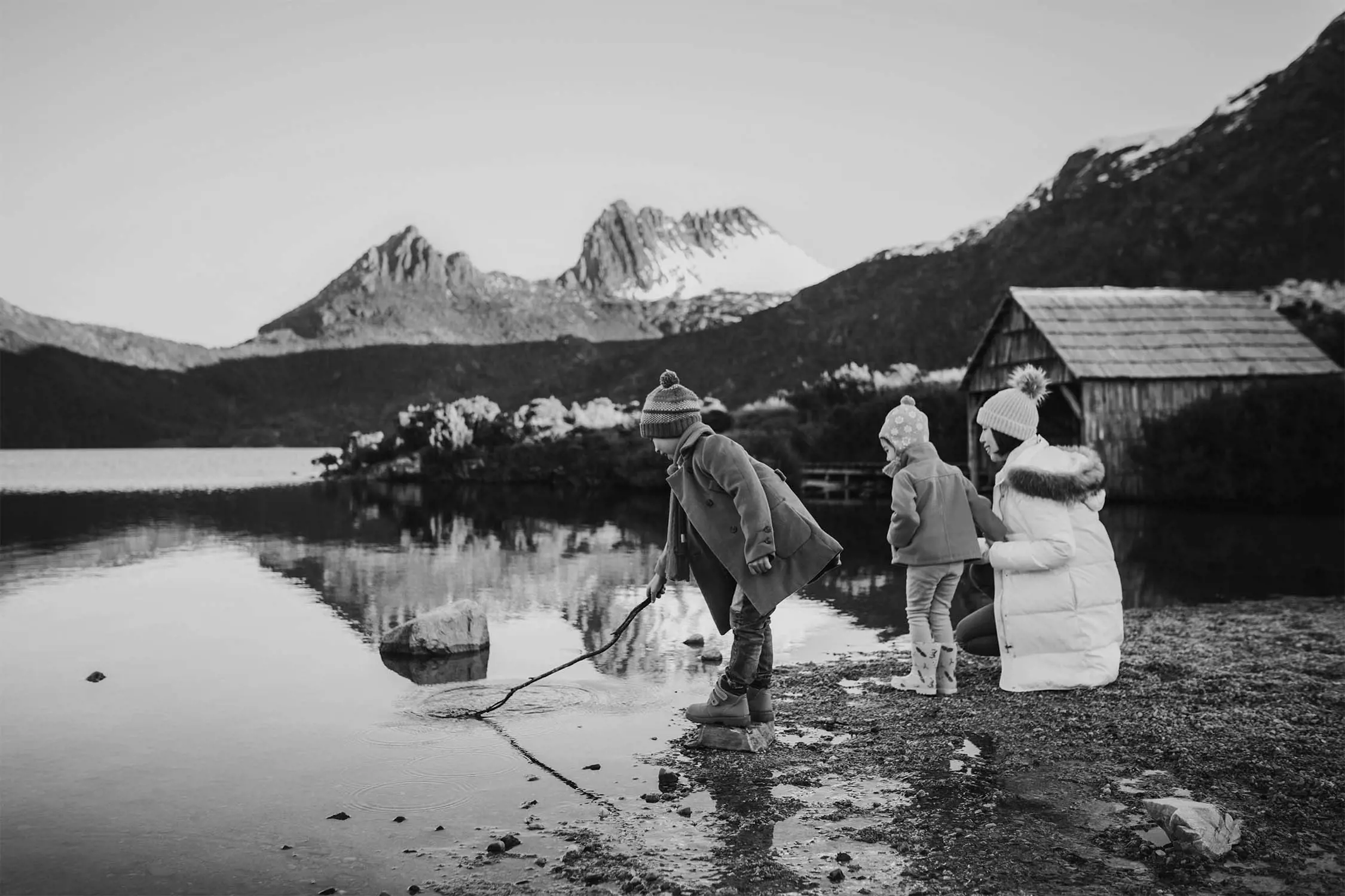 Children play near the water's edge of a lake, with mountains and a wooden cabin in the background.
