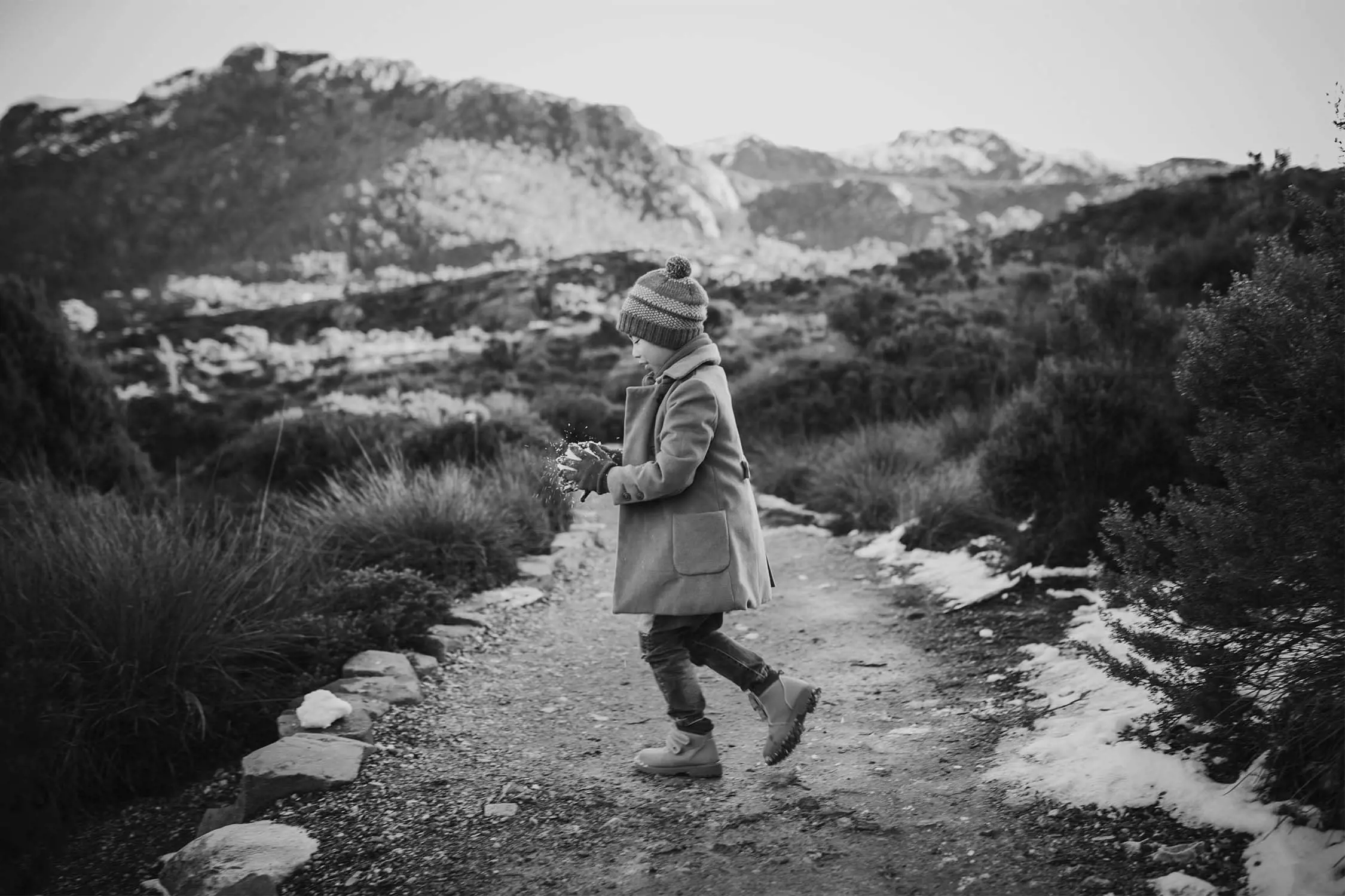 A child in winter attire walks along a path, tossing snowflakes in the air with mountains in the background.