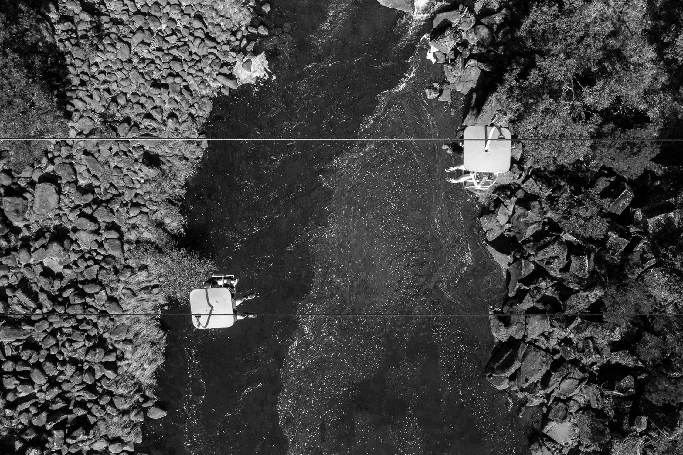 An aerial view captures two people on platforms above a river surrounded by rocky terrain
