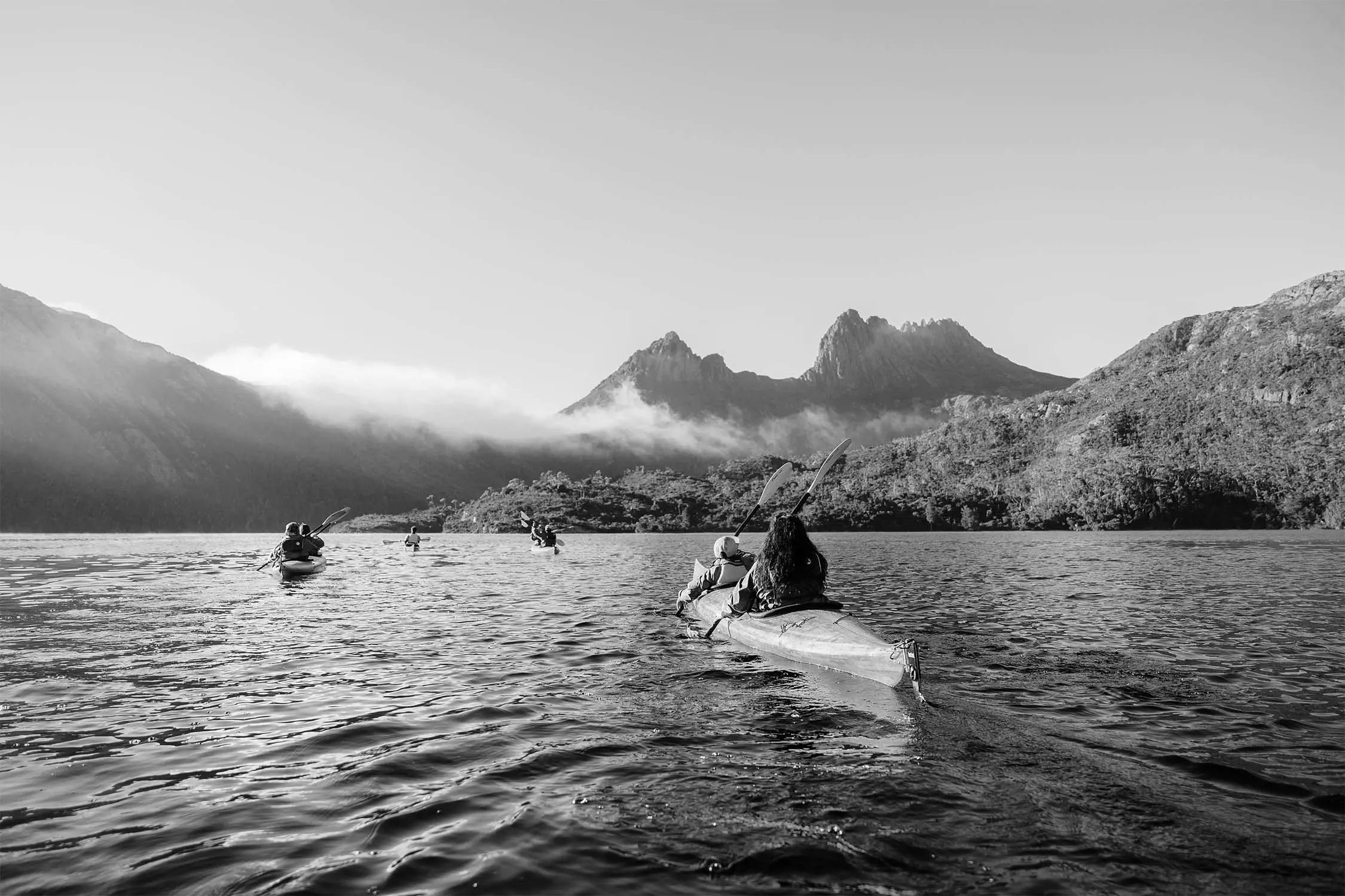 A group of kayakers paddle across a calm lake with mountainous terrain in the background.